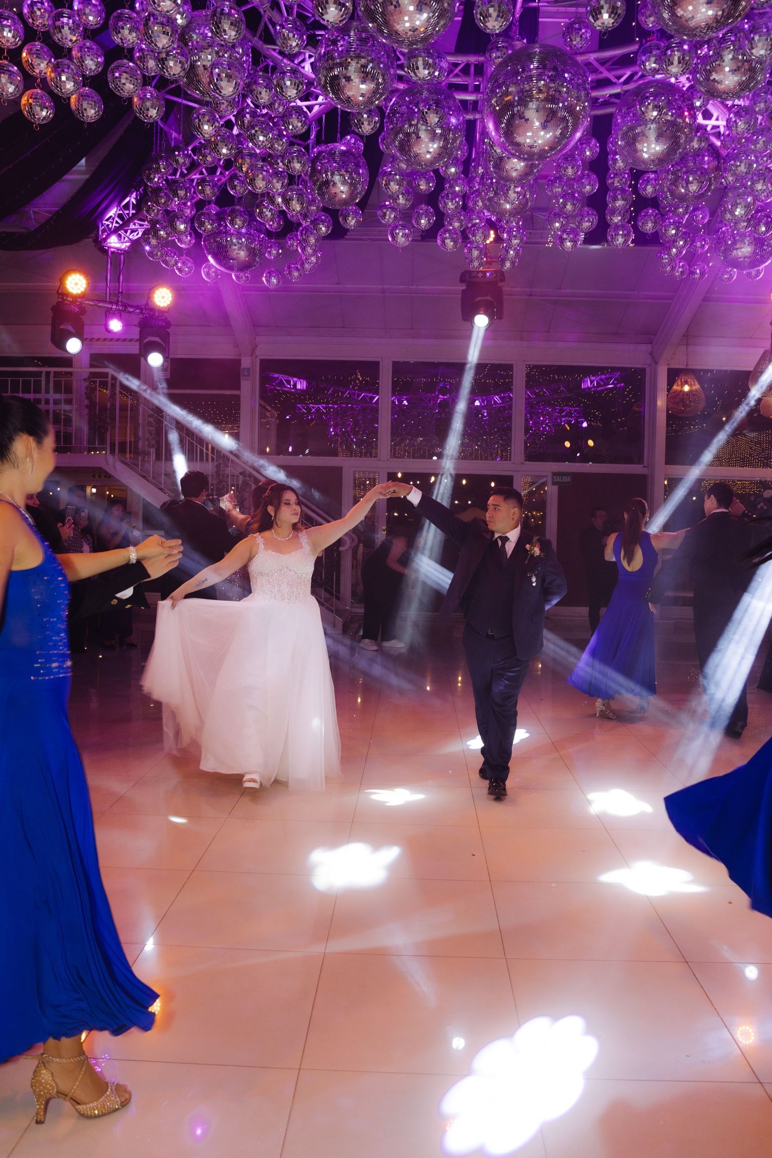 Bride and groom share first dance under disco balls and colorful spotlights on white dance floor at wedding reception.