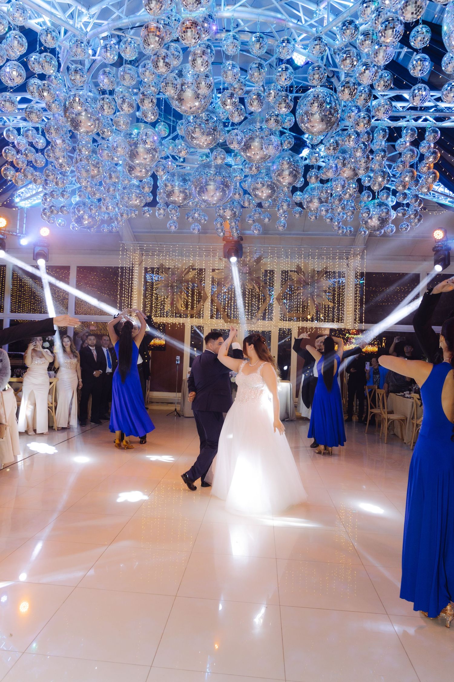 Bride and groom share first dance surrounded by bridesmaids in blue dresses under crystal ceiling decorations.
