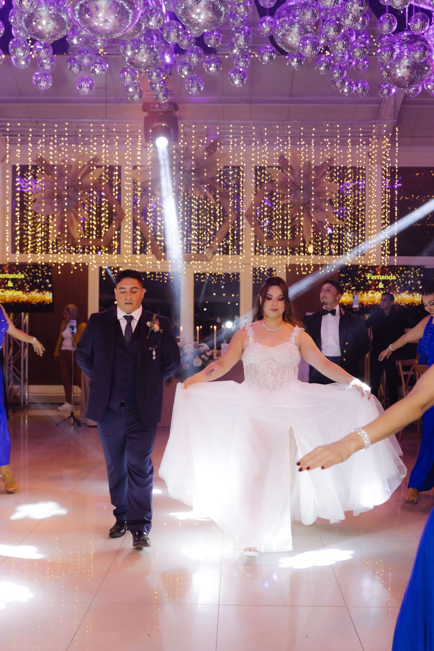 Bride in white gown and groom in navy suit walk onto illuminated wedding reception dance floor.