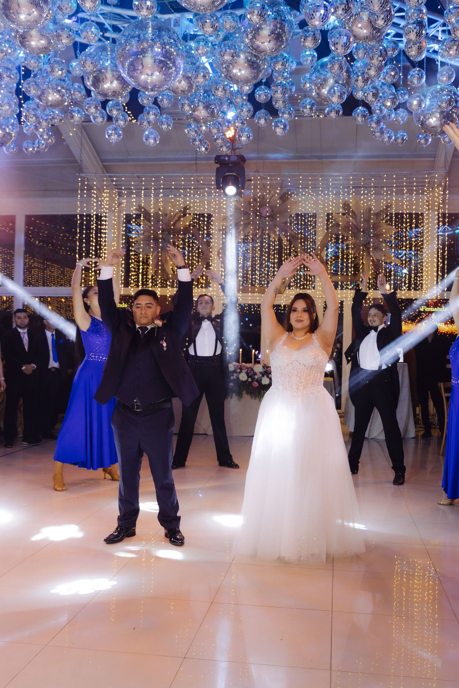 Bride and groom raise arms together during choreographed first dance with wedding party at reception.