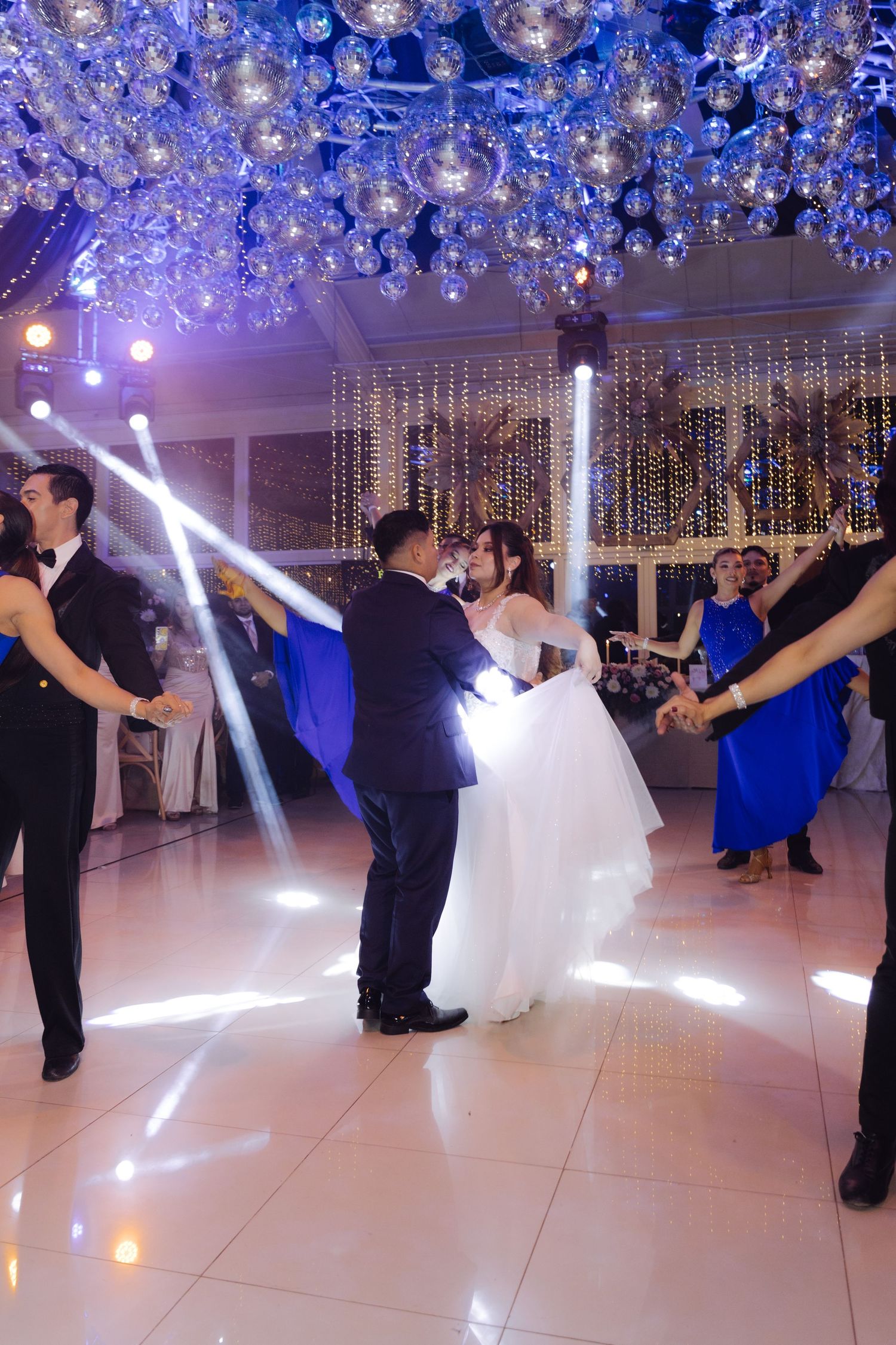 Bride and groom dance romantically surrounded by wedding party under sparkling blue-lit crystal ceiling.