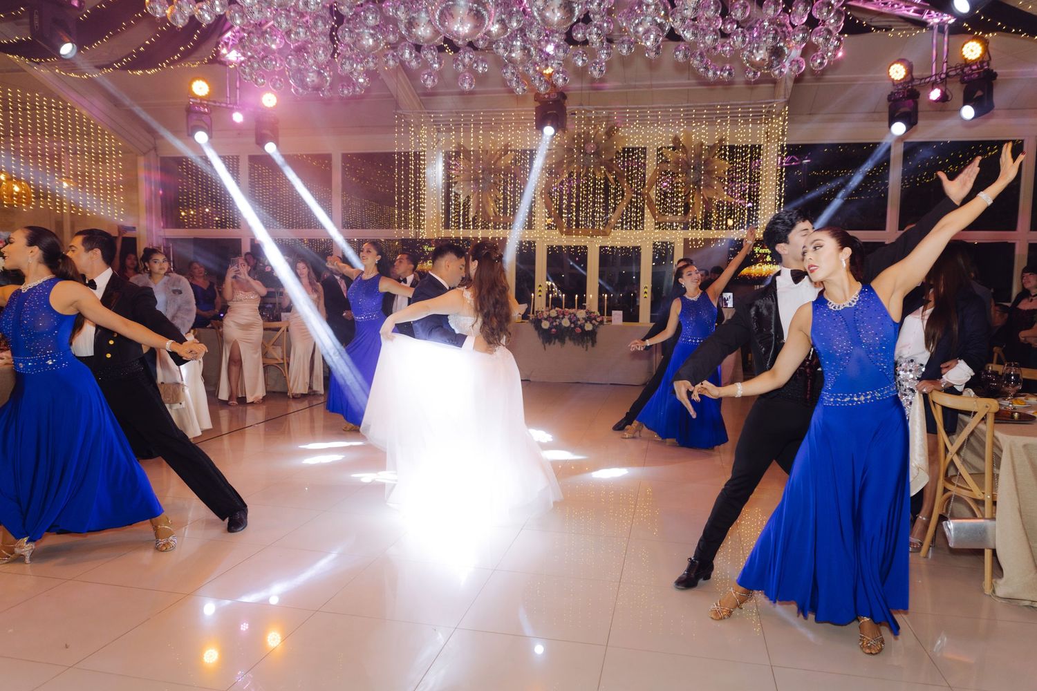 Professional dancers in blue ballroom gowns perform alongside bride and groom at wedding reception.