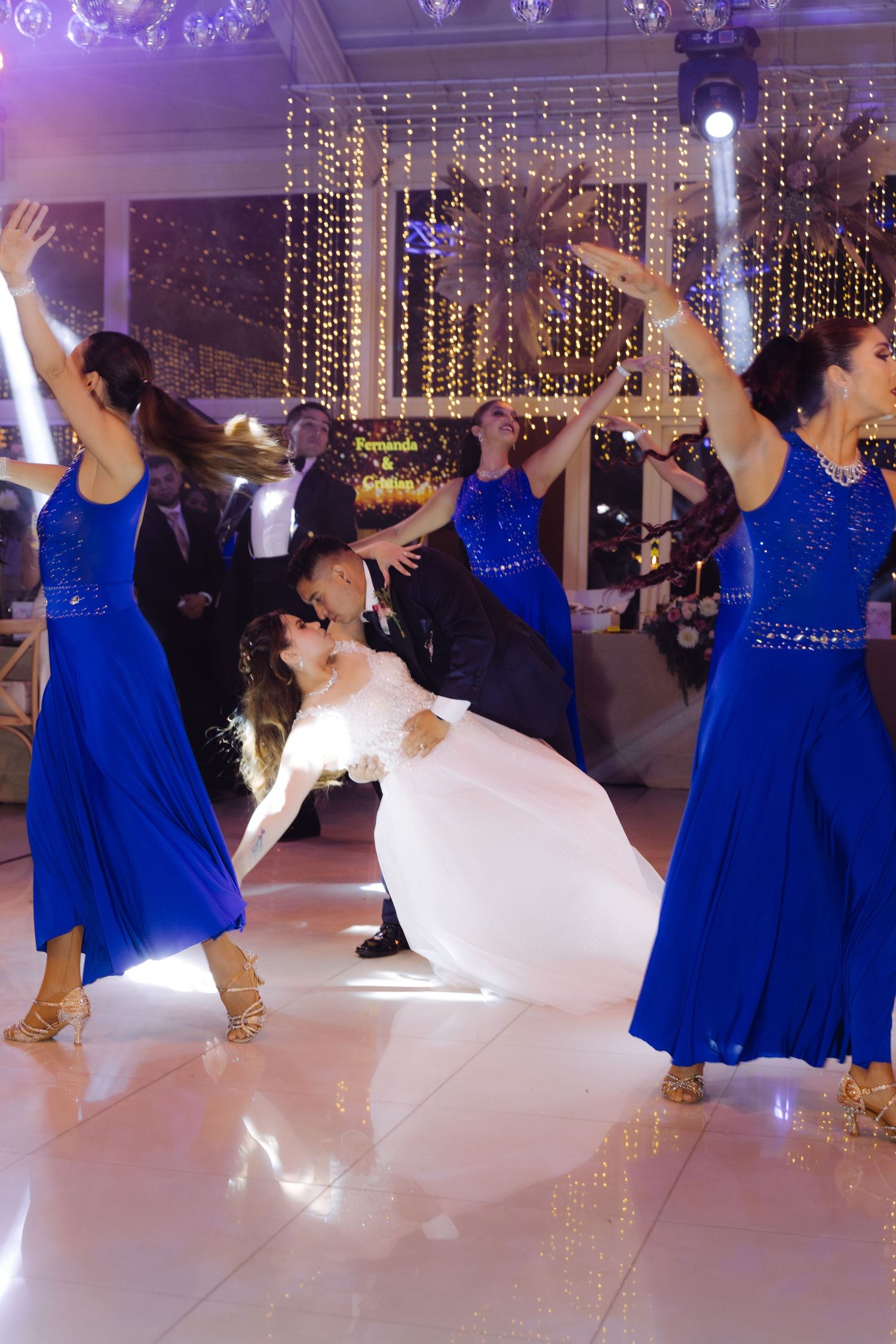 Groom dips bride dramatically during first dance while professional dancers in blue dresses perform around them.