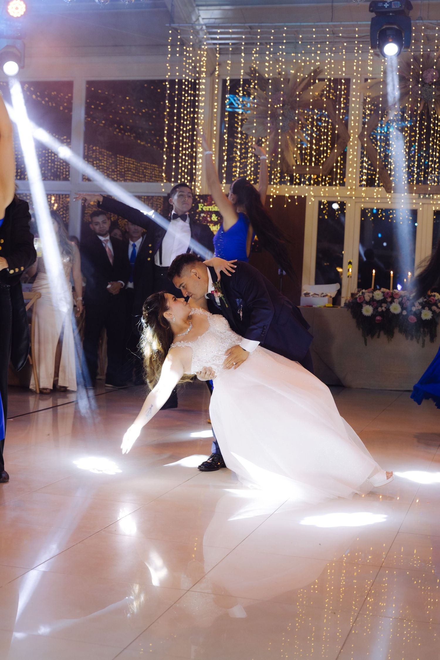 Groom dips bride in white wedding gown under spotlight beams at elegant reception venue.