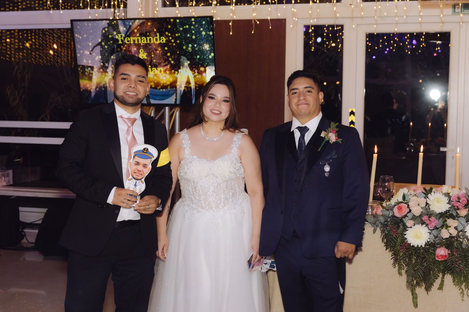 Bride in lace wedding gown poses with two guests holding a decorative photo prop at reception.