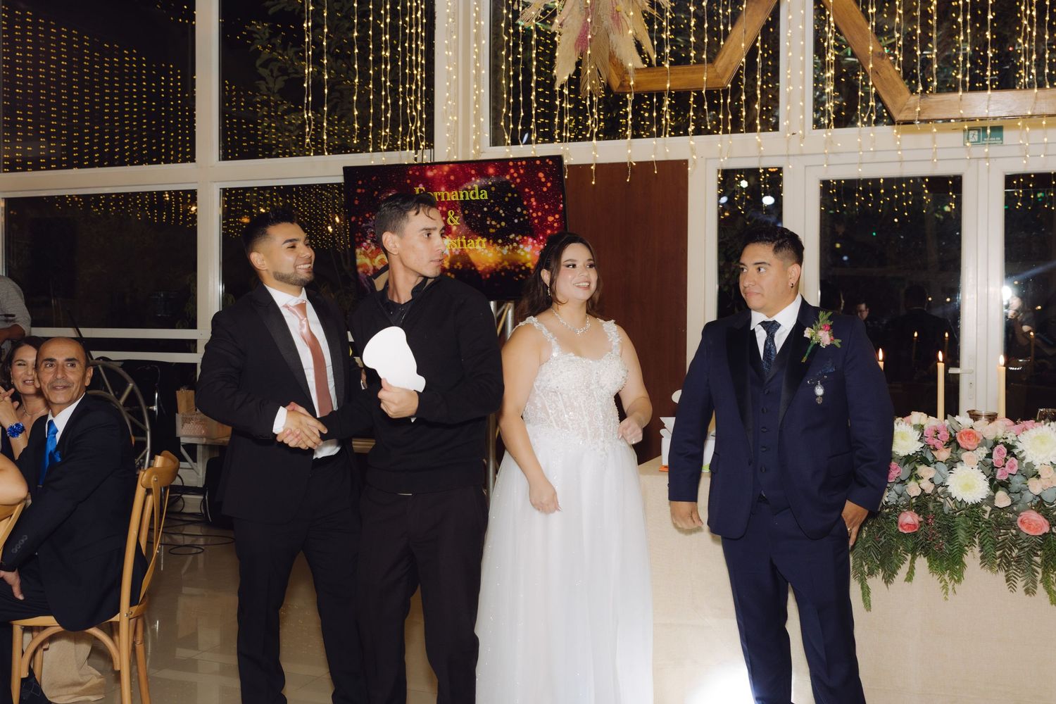 Bride in white gown stands with wedding guests near floral arrangements at indoor reception venue.