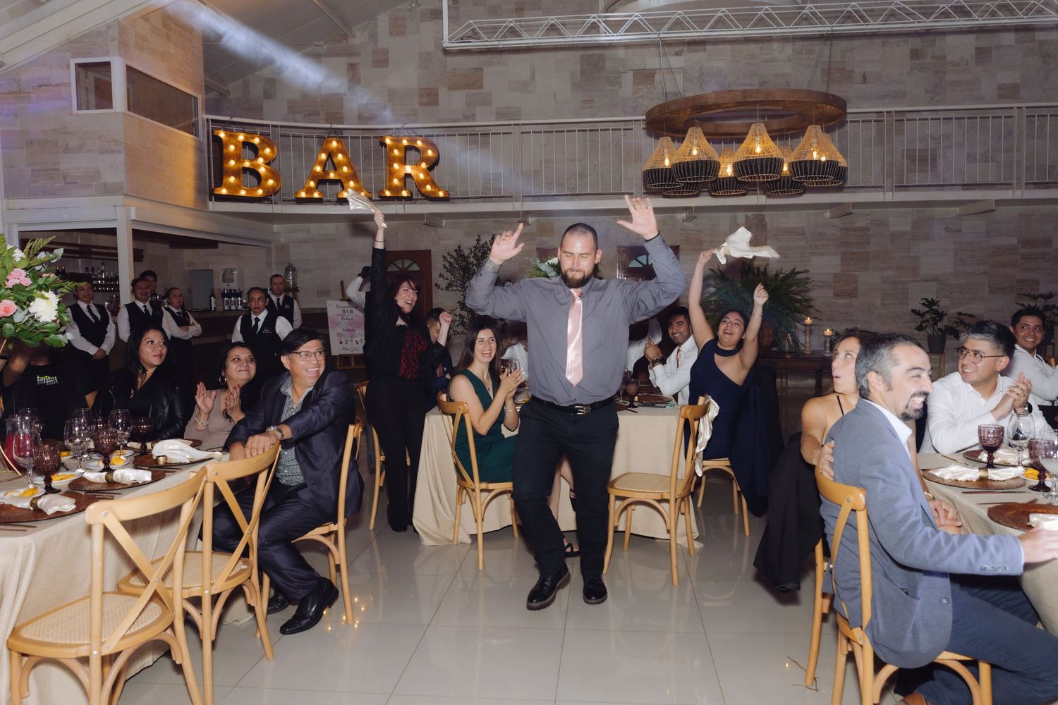 Man dancing energetically at wedding reception with guests seated at decorated tables and BAR sign in background.