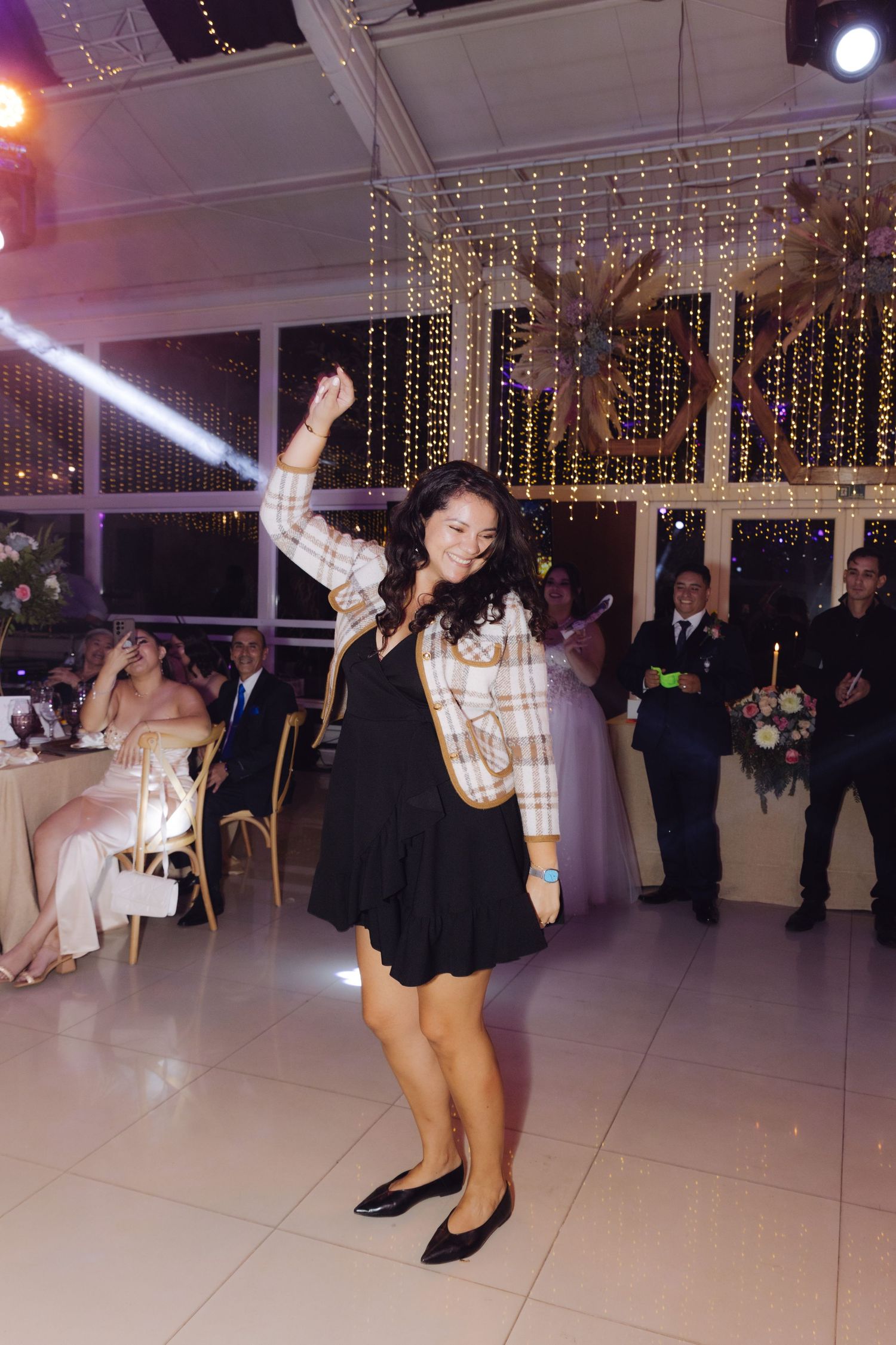 Young woman dancing joyfully at wedding reception with one arm raised, wearing black dress and plaid jacket.