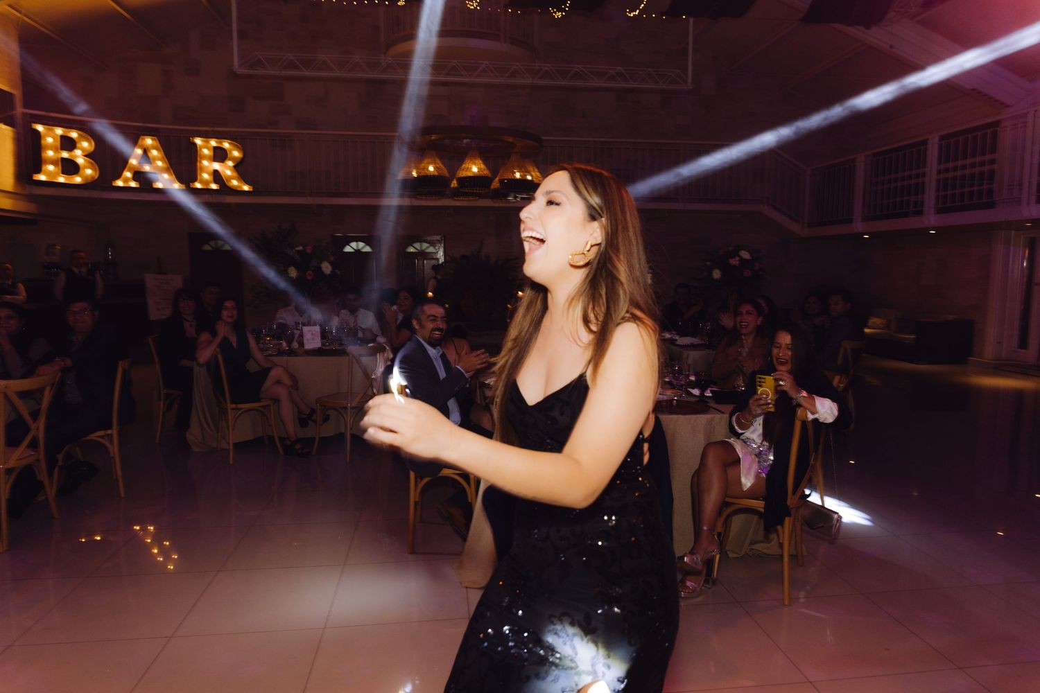 Woman in black sequin dress dancing and celebrating at elegant wedding reception with BAR sign behind her.