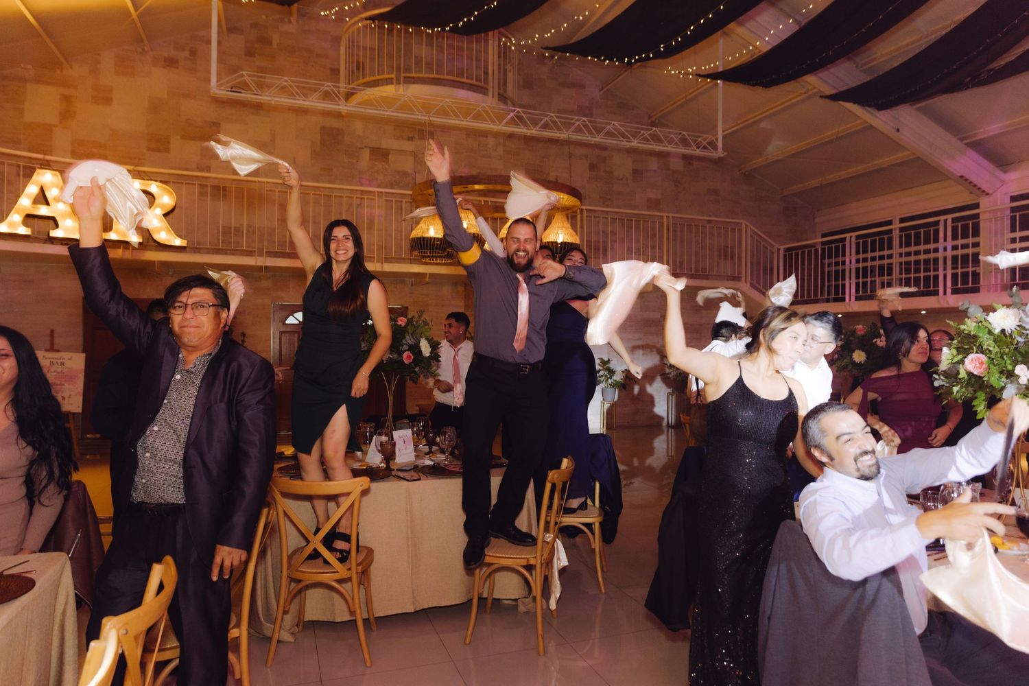 Wedding reception guests enthusiastically waving white napkins while standing on chairs at decorated venue.