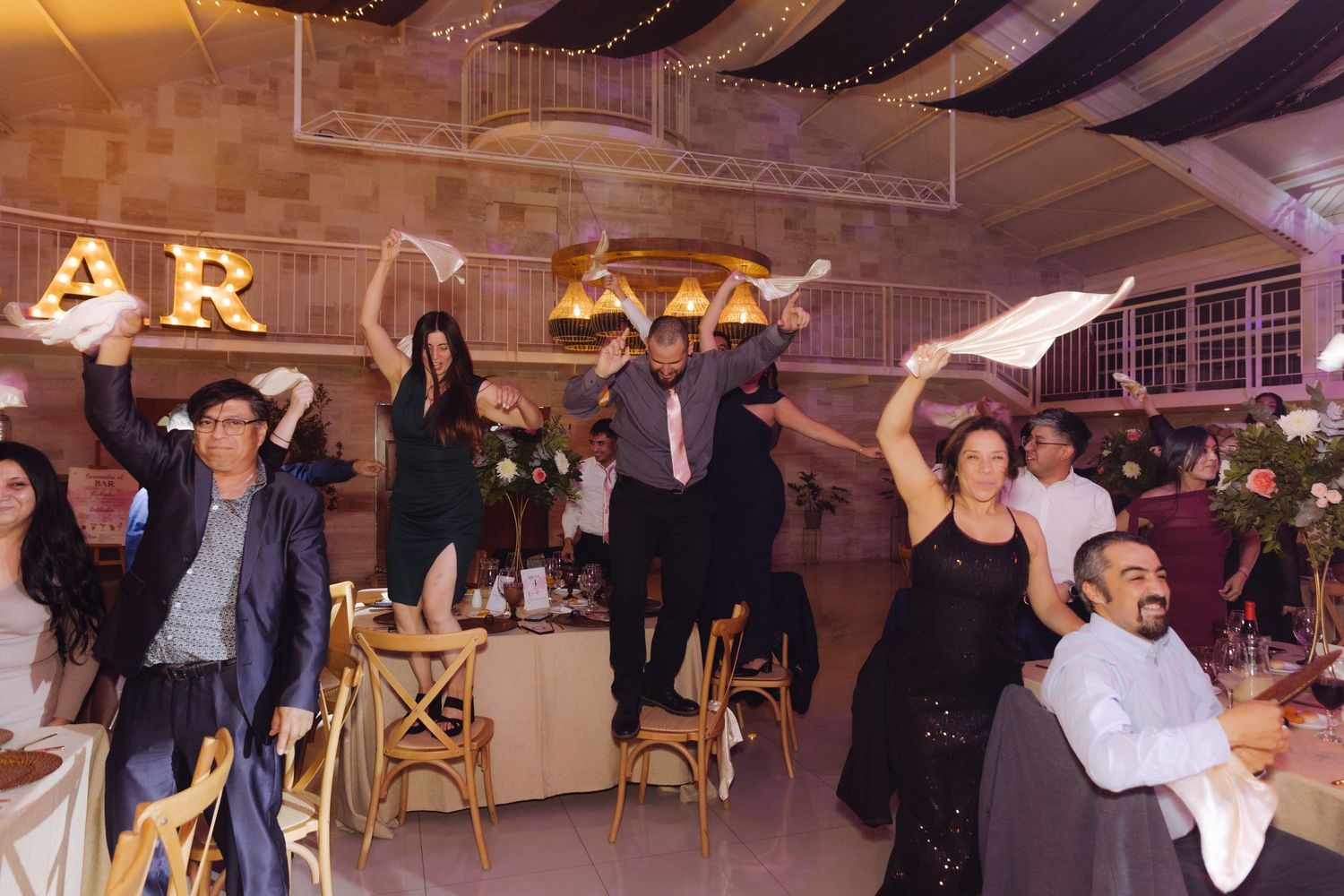 Excited wedding guests standing on chairs waving white napkins in celebration at festive reception venue.