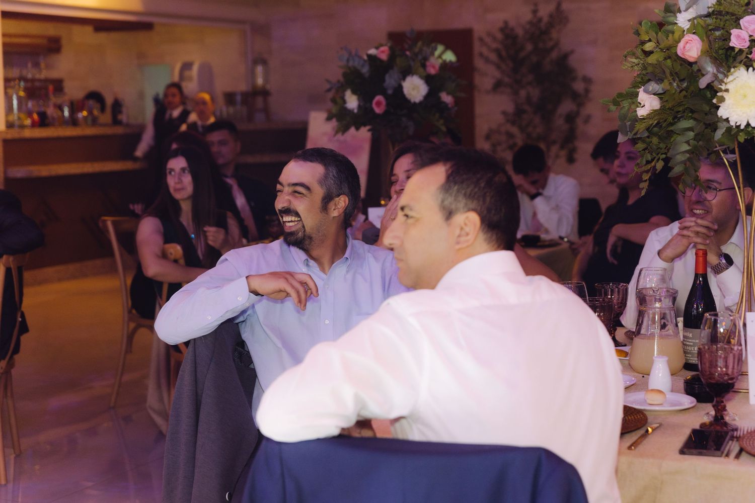 Two men laughing and conversing at wedding reception table with drinks and floral decorations nearby.
