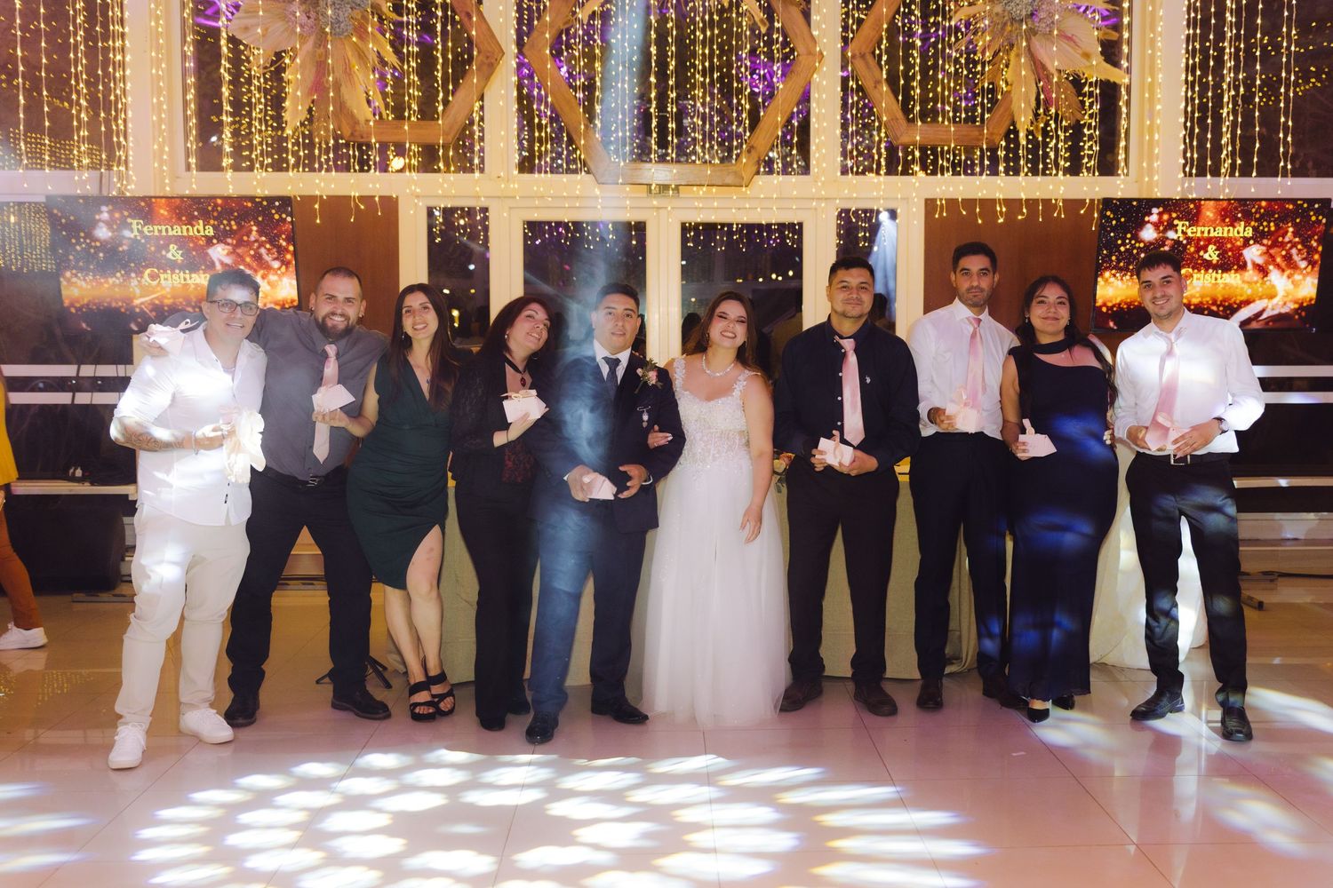 Wedding party group photo on dance floor with bride in white gown surrounded by formally dressed guests.