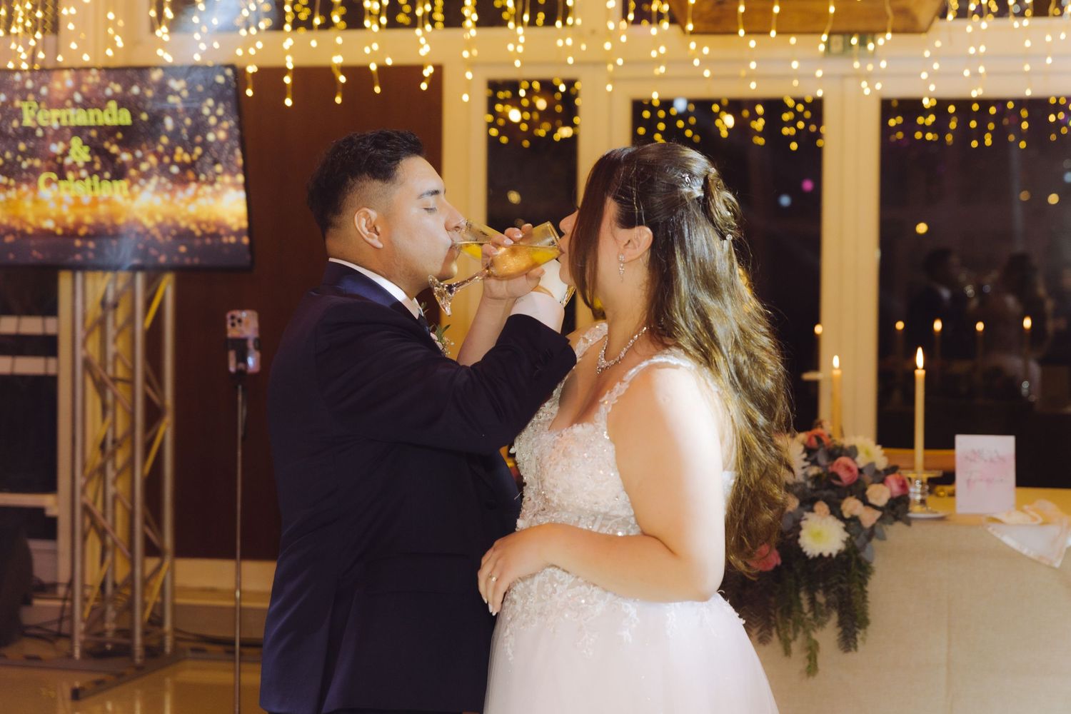 Bride and groom sharing a romantic champagne toast at their wedding reception with fairy lights behind them.