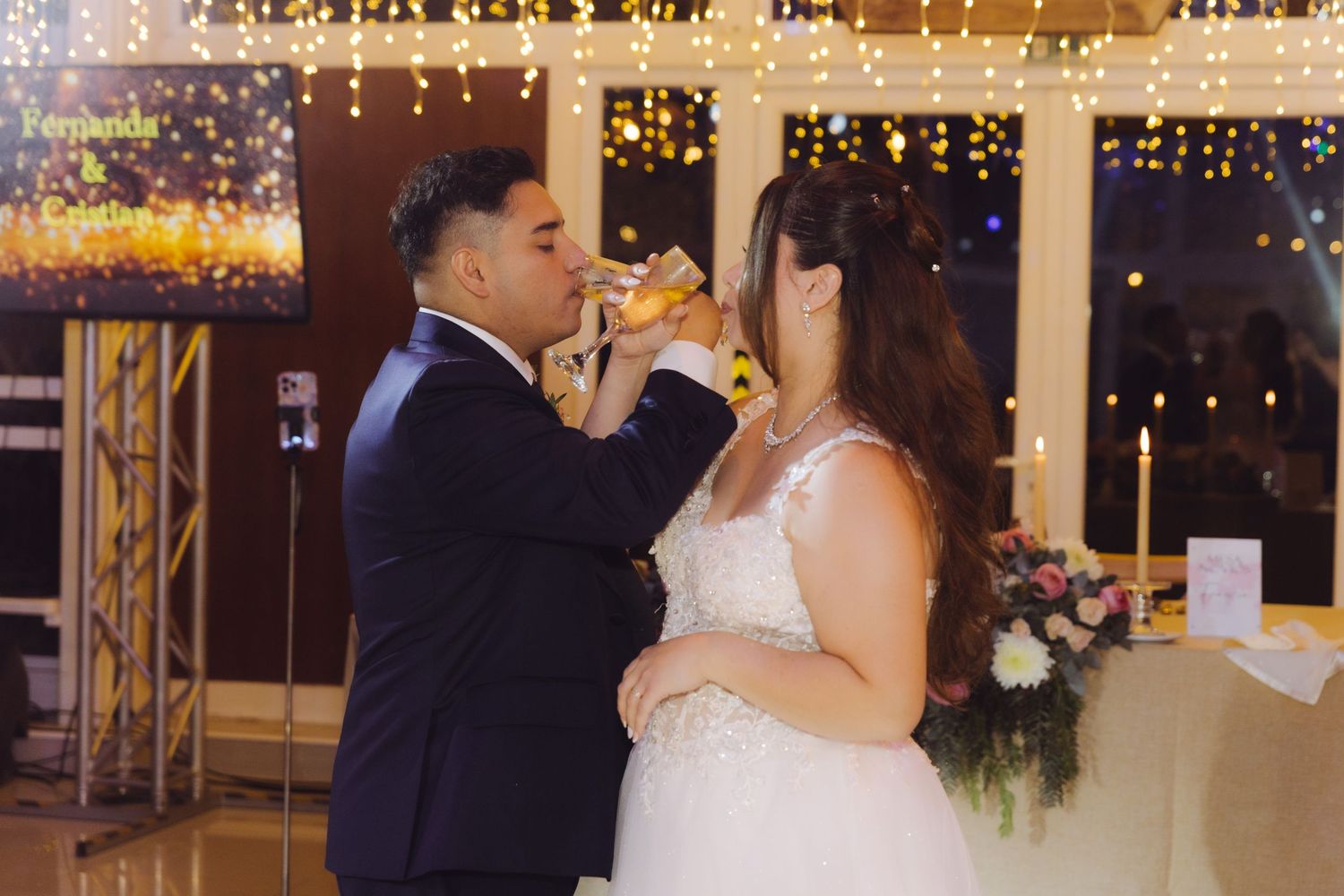 Bride and groom sharing a toast with champagne glasses at their elegant wedding reception with fairy lights.