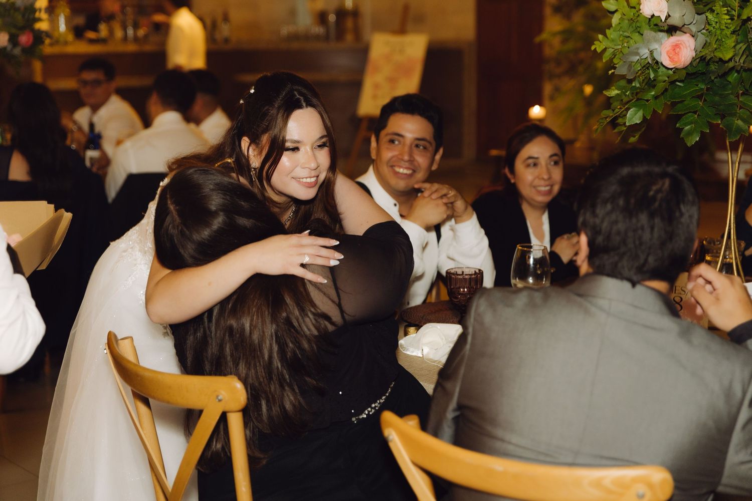 Bride in white gown warmly embracing a guest in black dress at the wedding reception.