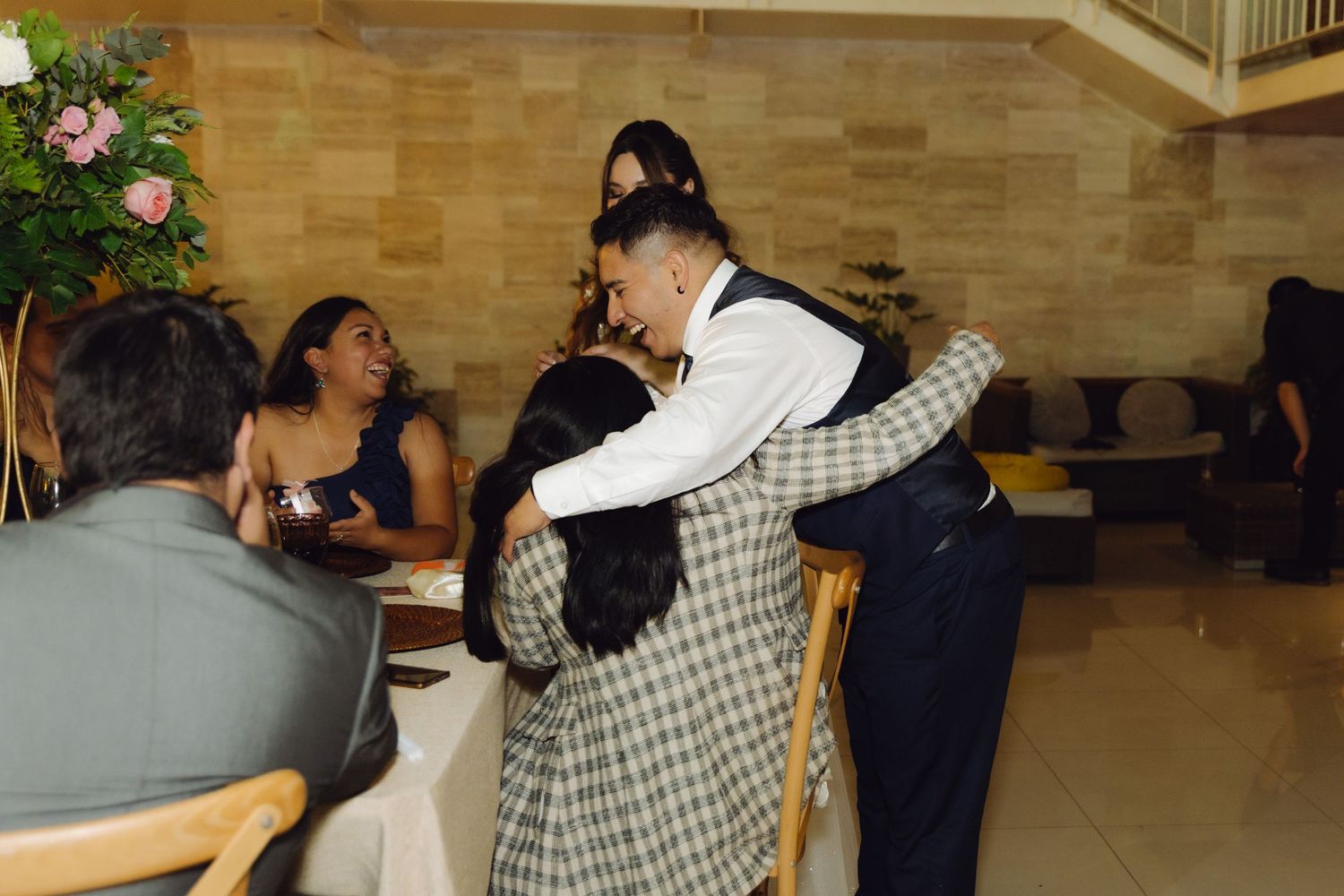 Groom in vest hugging a wedding guest near elegantly decorated tables at the reception.