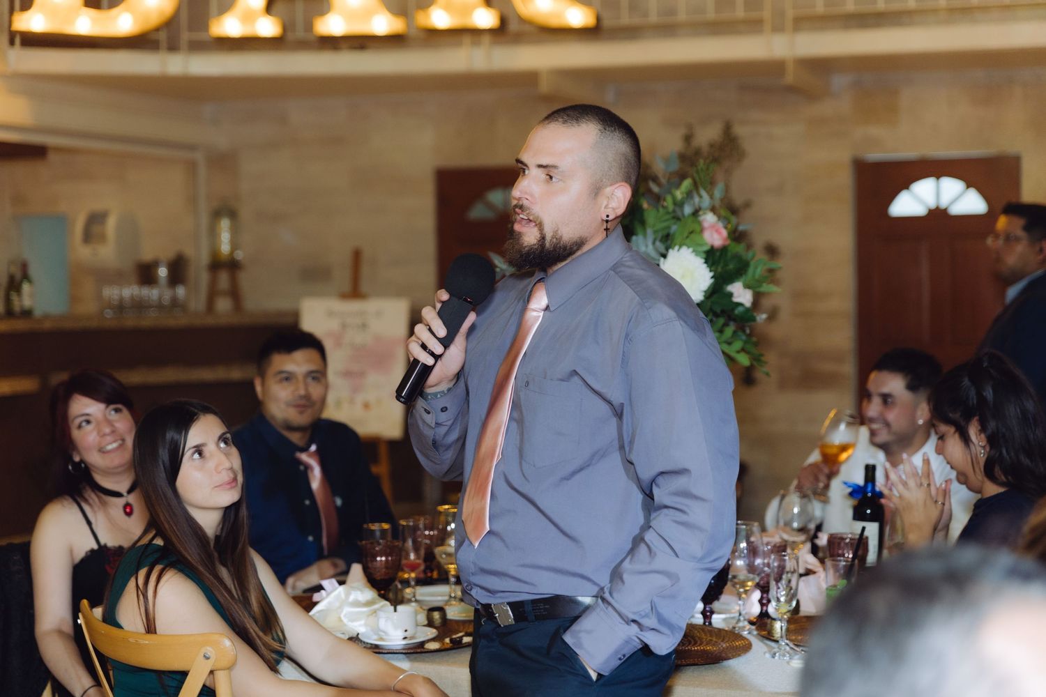 Man in blue shirt giving a heartfelt speech with microphone at a wedding reception.