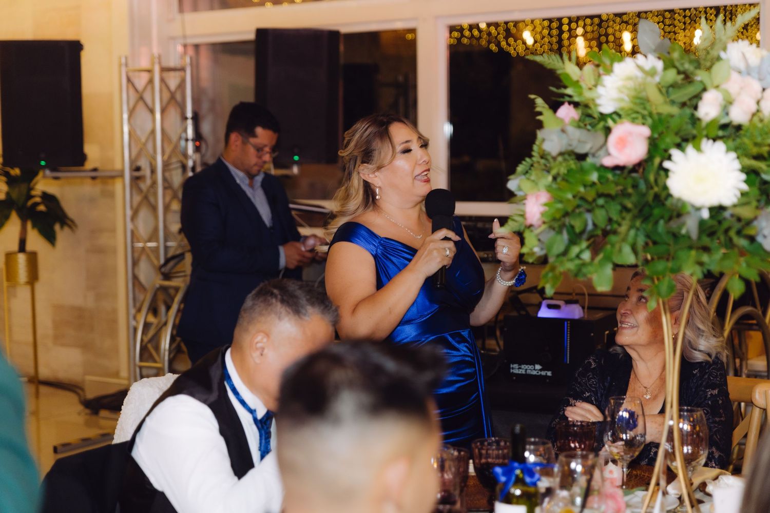Elegant woman in blue dress speaking into microphone at a wedding reception with floral decor.