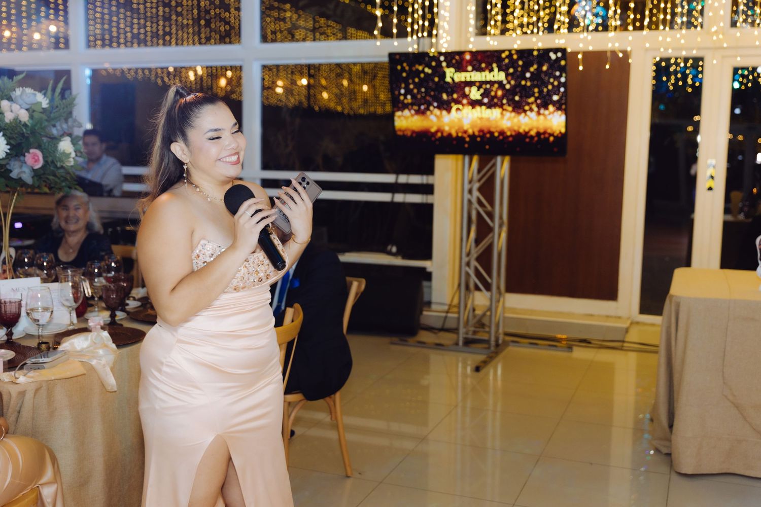 Woman in elegant gown speaking into microphone at wedding reception with festive lights in background.