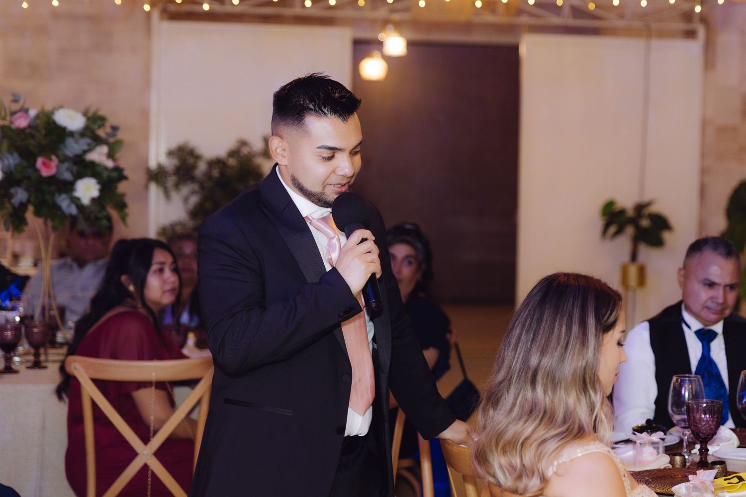 Groom in black suit delivering heartfelt speech into microphone at elegant wedding reception dinner.