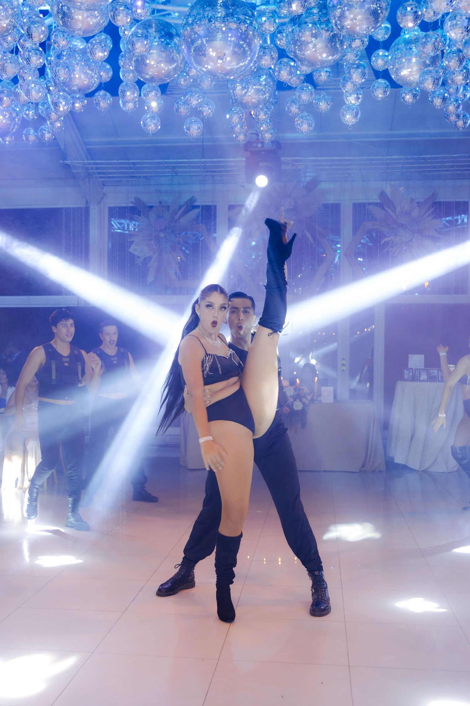 Professional dance couple performing dramatic lift routine under sparkling balloon ceiling with blue stage lighting.