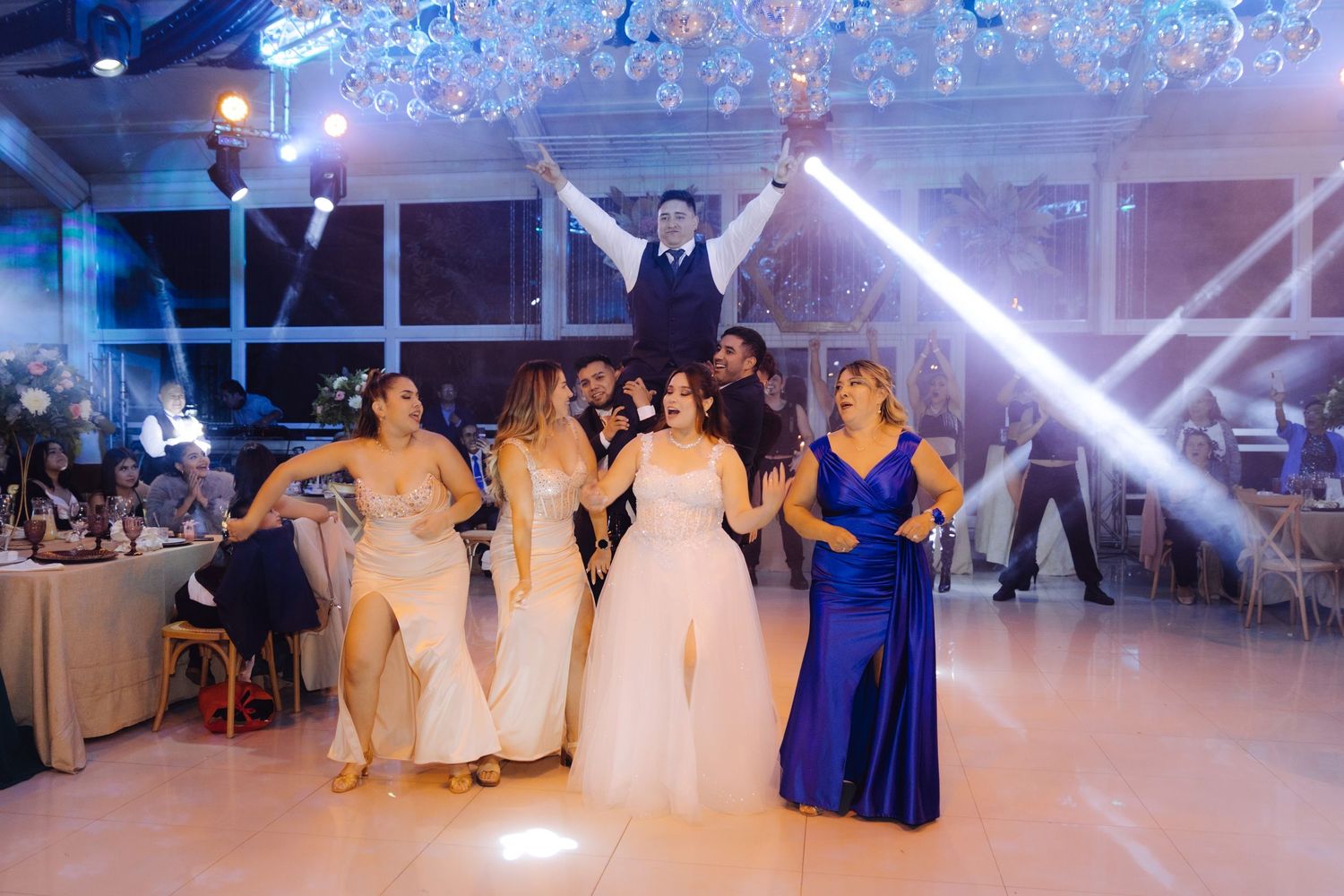 Bride and bridesmaids in colorful gowns dancing joyfully on illuminated wedding reception dance floor.