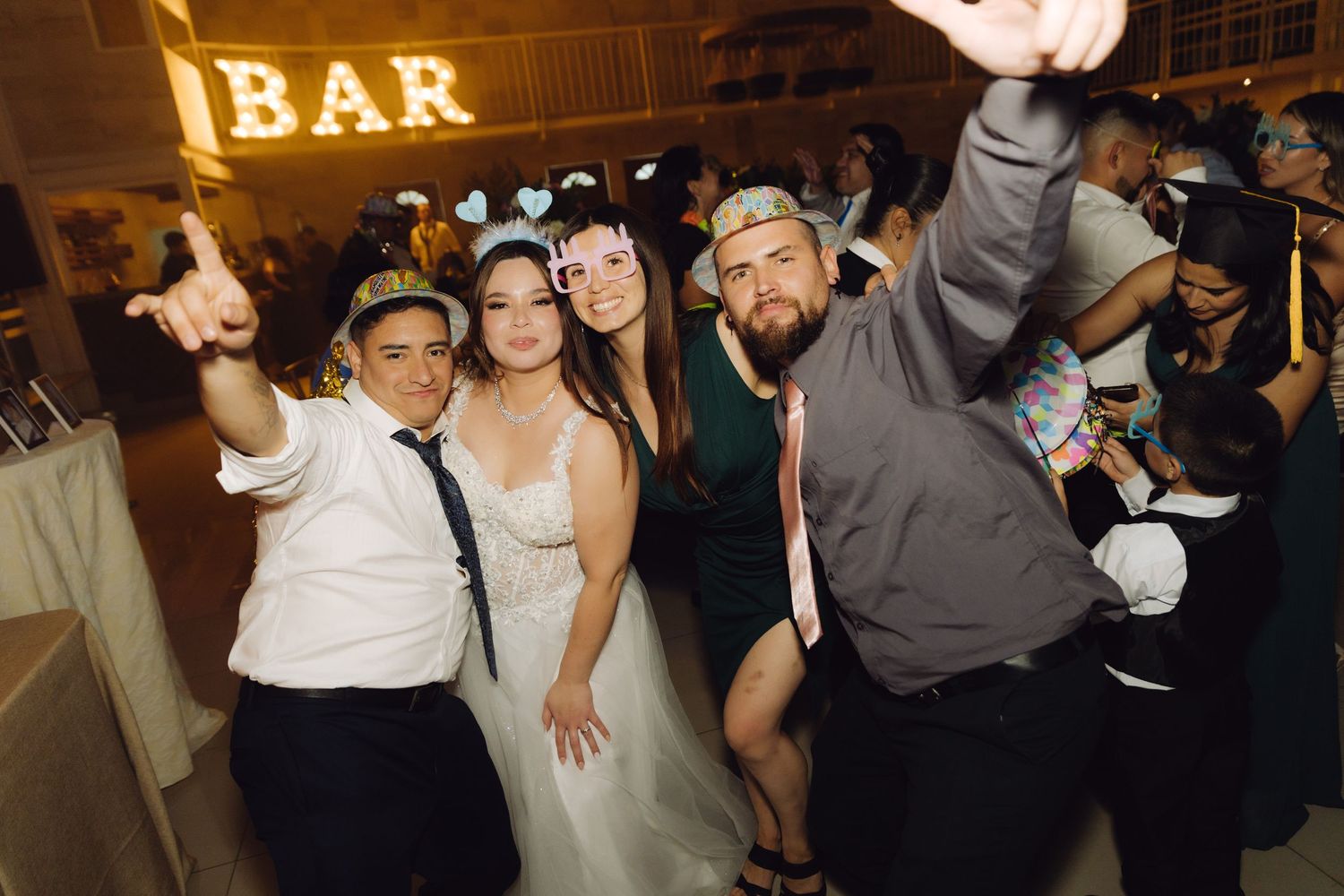 Bride and wedding guests taking fun selfie wearing party hats near illuminated BAR sign at reception.