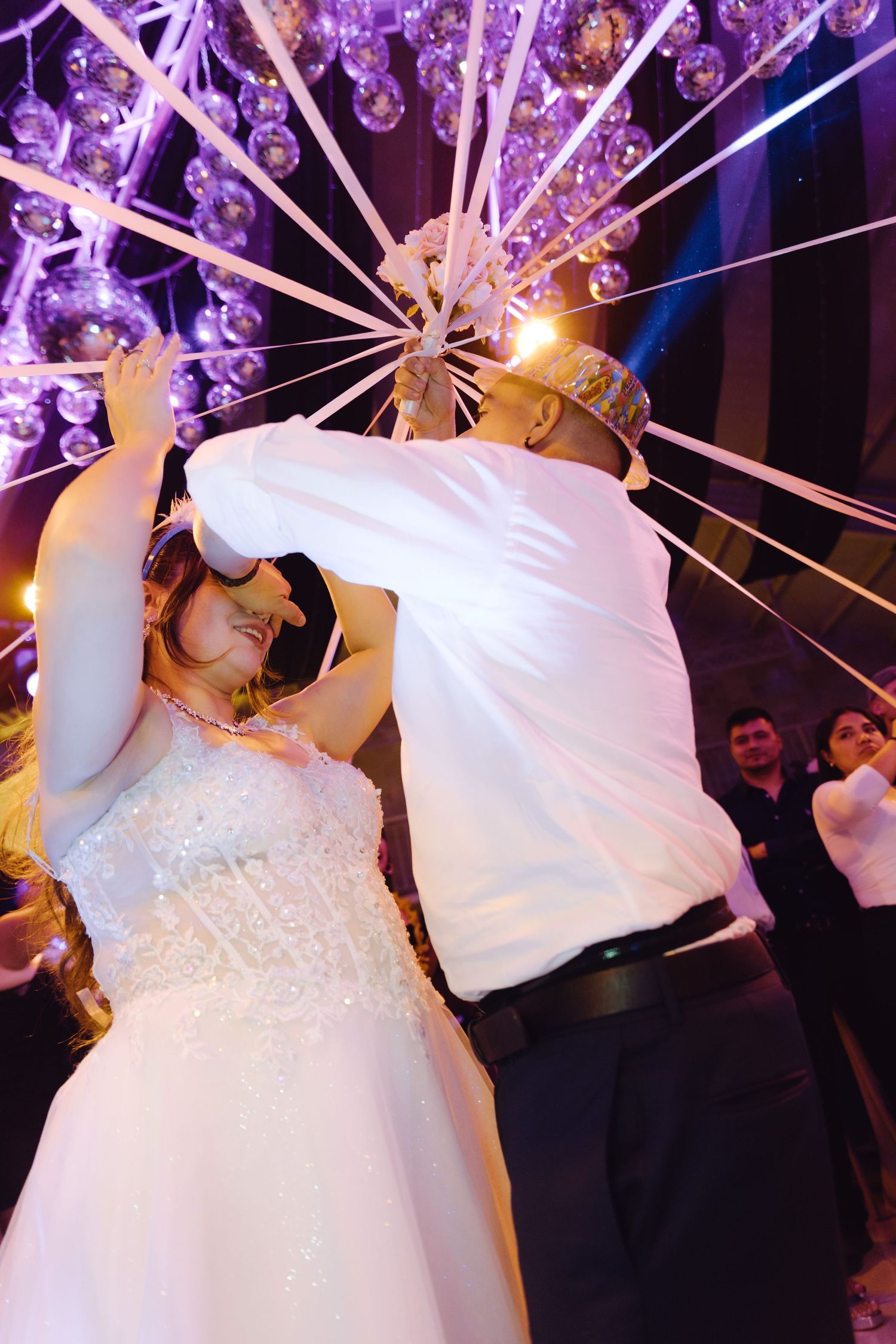 Bride and groom sharing blindfolded first dance under sparkling chandelier at wedding reception.
