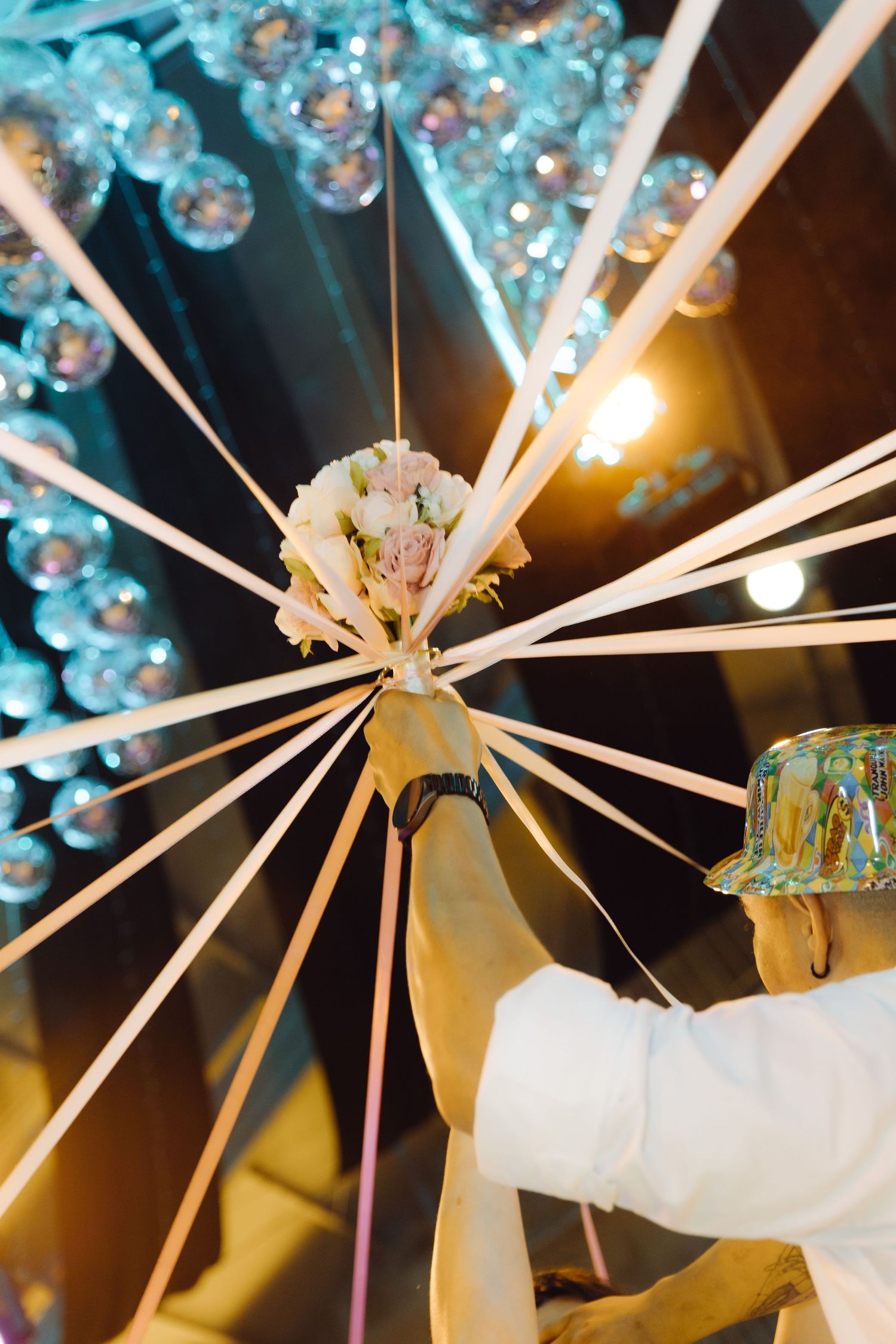 Groom holding wedding bouquet with ribbons fanning out under decorative crystal ball ceiling at reception.