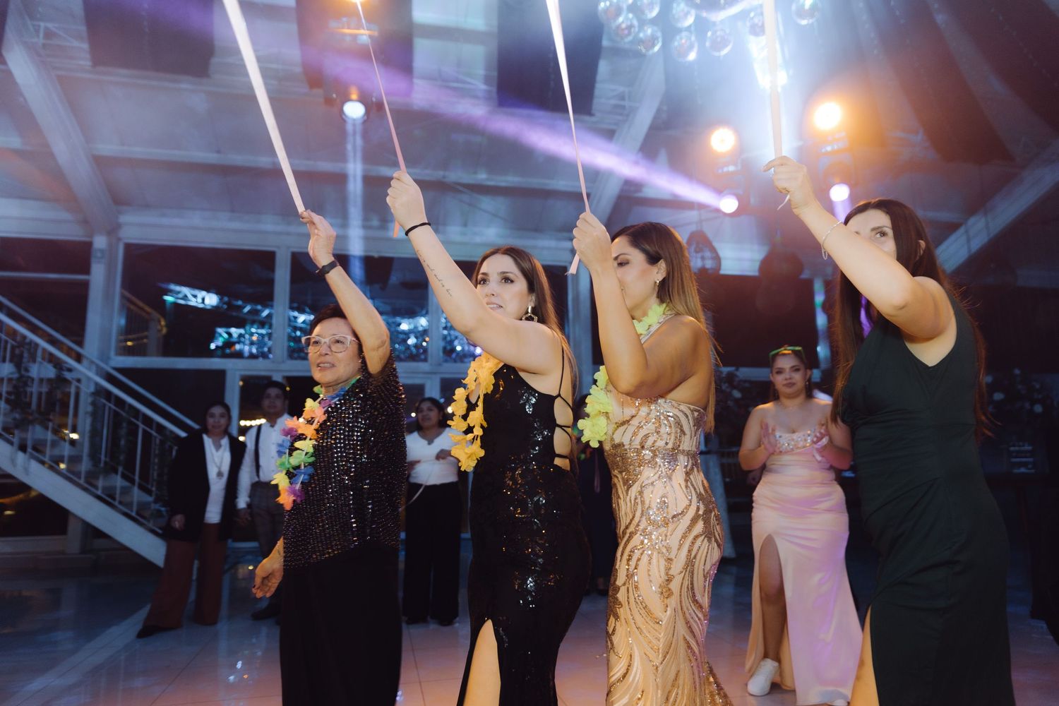 Female wedding guests holding ribbon wands on dance floor during lively evening reception celebration.