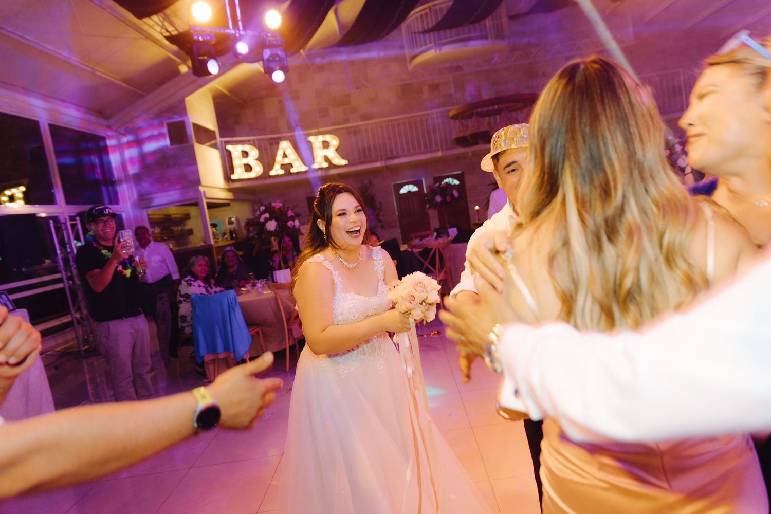 Joyful bride in white gown dancing and laughing with guests near illuminated BAR sign at reception.