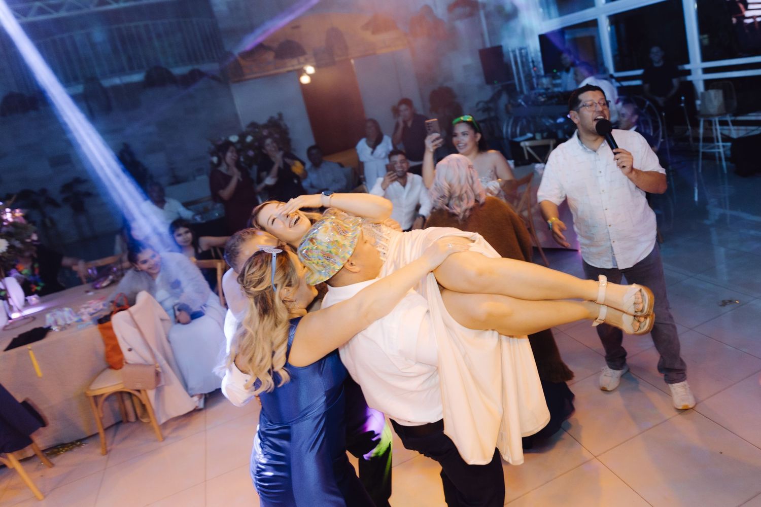 Groom lifting bride on wedding dance floor while guests cheer and MC performs at reception.