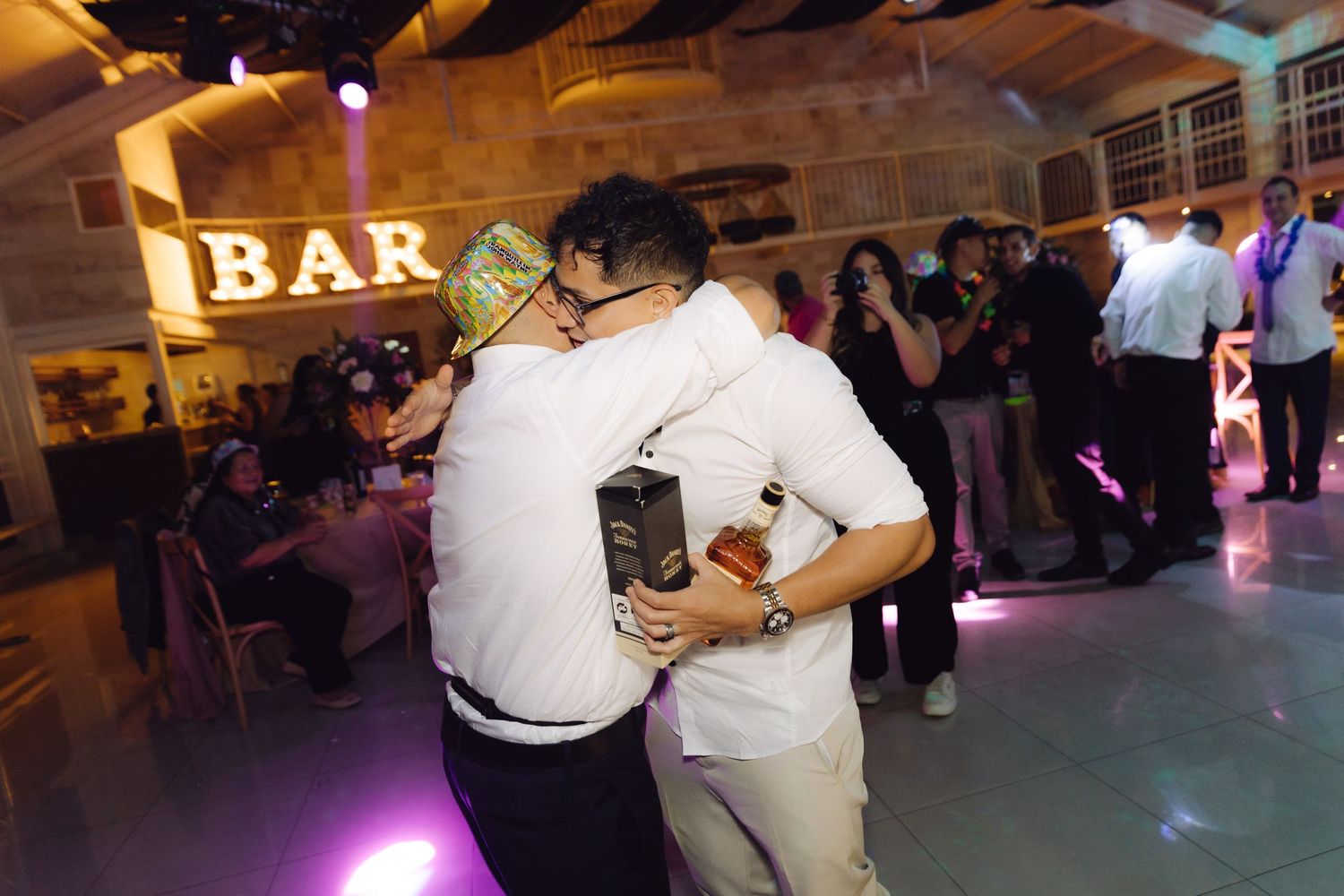 Two guests sharing a warm hug on the dance floor near a glowing BAR sign at a wedding.