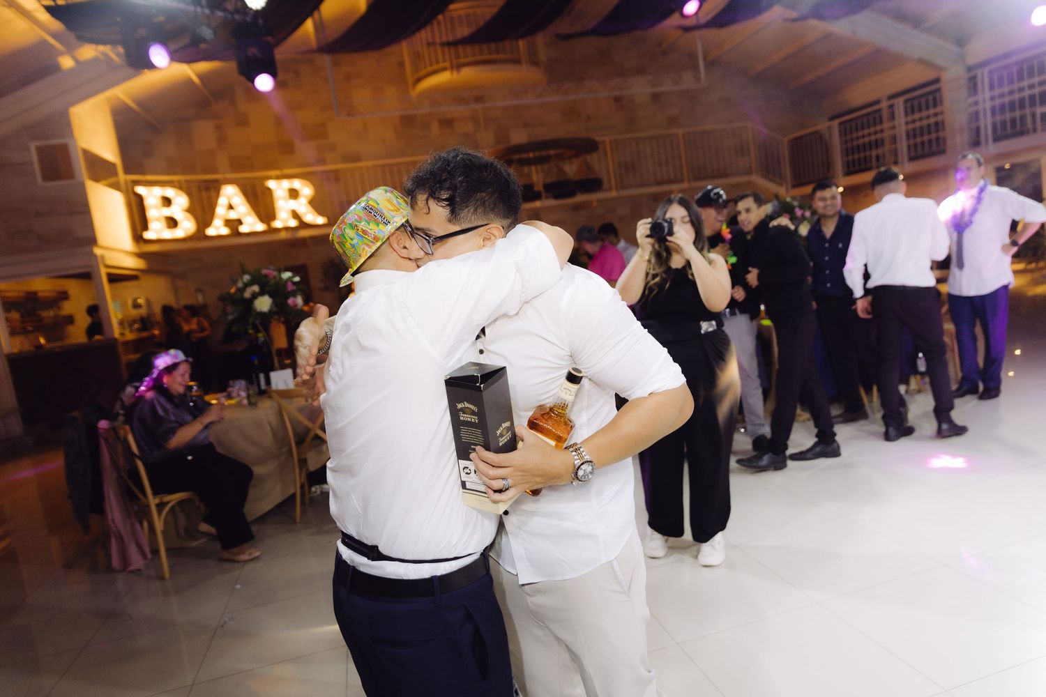 Two guests embracing on the dance floor near an illuminated BAR sign at a wedding reception.