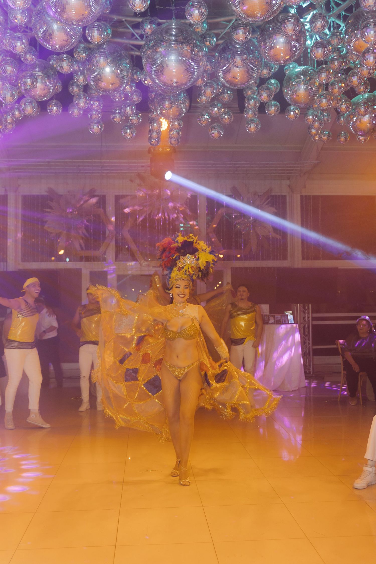 Colorful carnival dancer in yellow feathered costume performing under disco ball ceiling at a gala event.