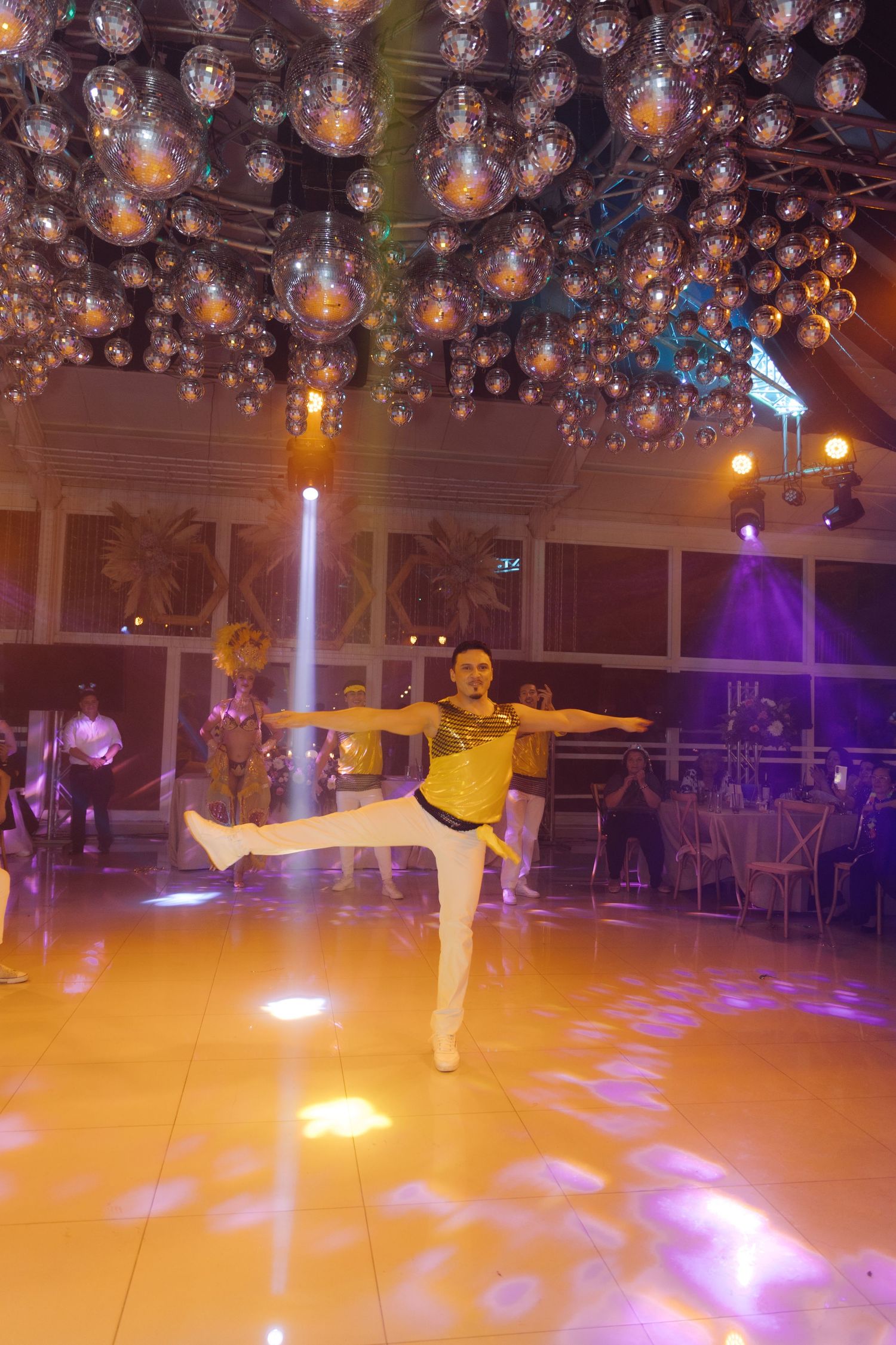 Male performer in gold costume doing a high kick under a sparkling disco ball ceiling at a gala.