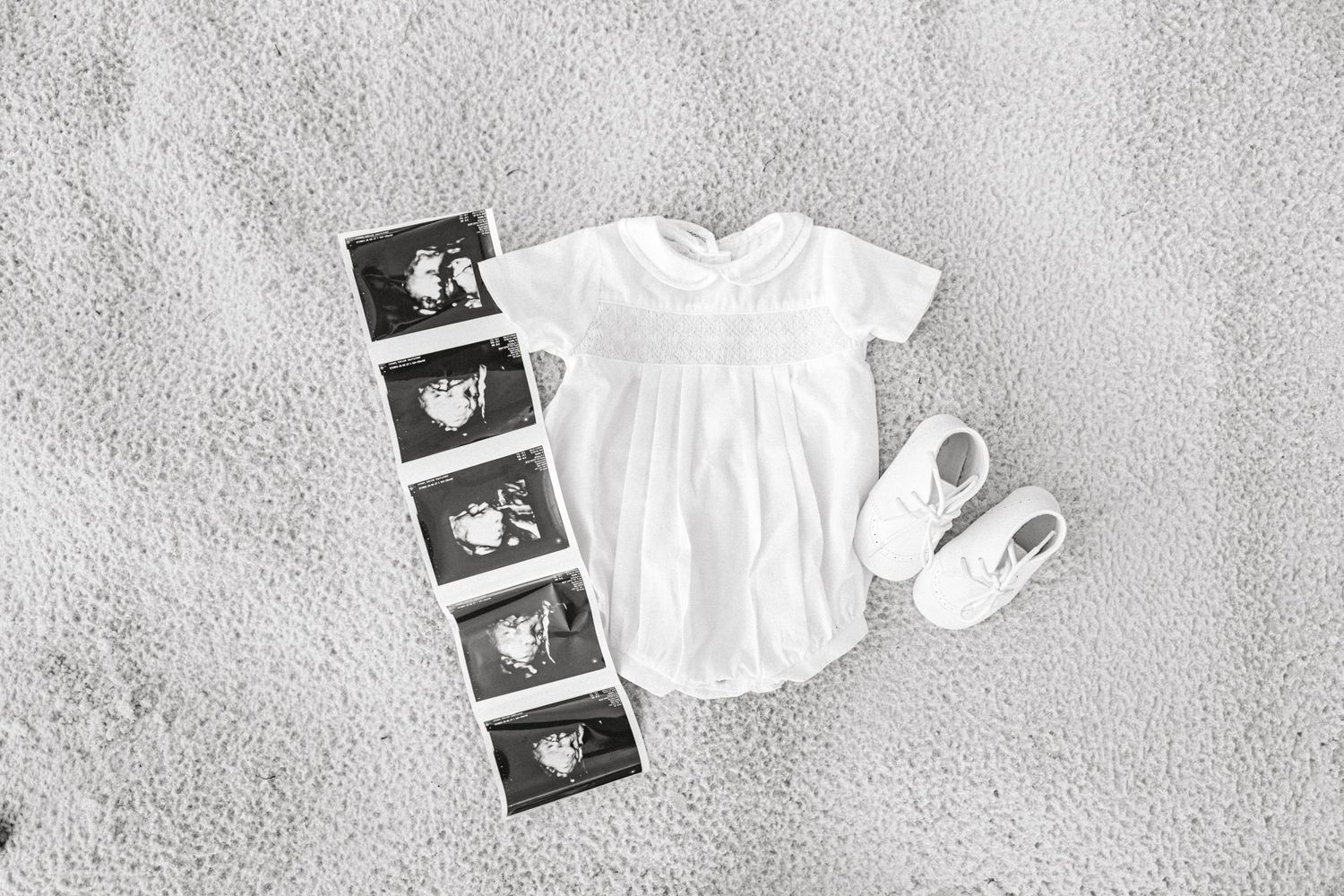 Black and white flat lay of baby onesie, ultrasound photos, and white booties on soft carpet.
