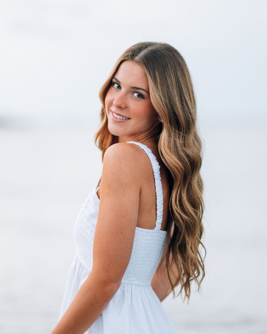 Young woman with long wavy brown hair smiles over shoulder in white sundress on a beach.