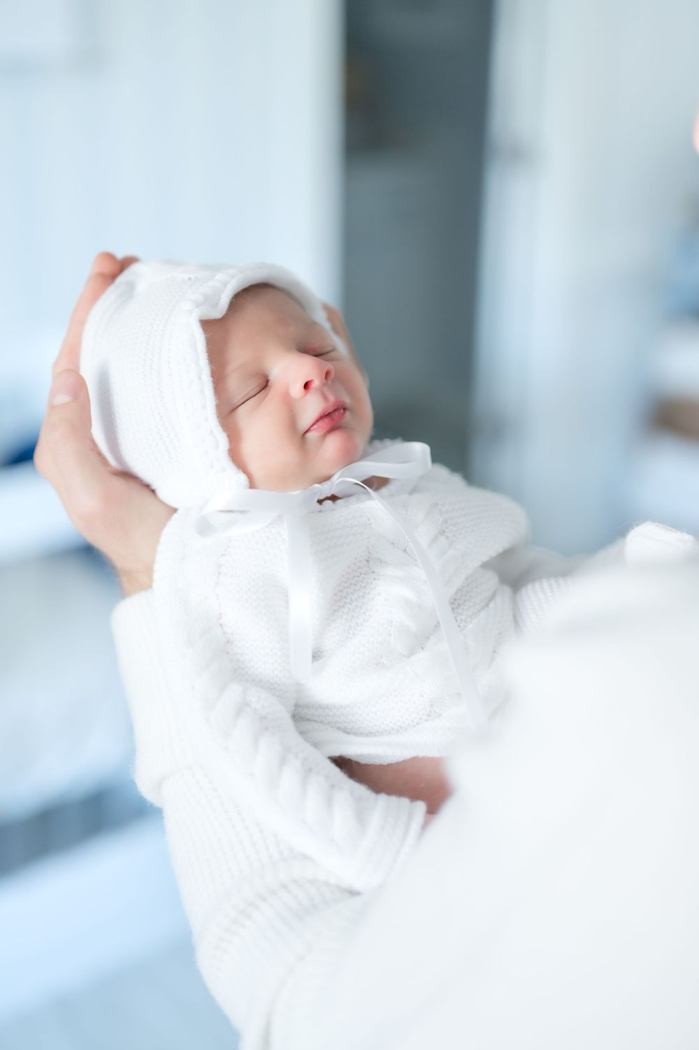 A sleeping newborn baby wrapped in soft white blankets and wearing a white knit cap rests peacefully.
