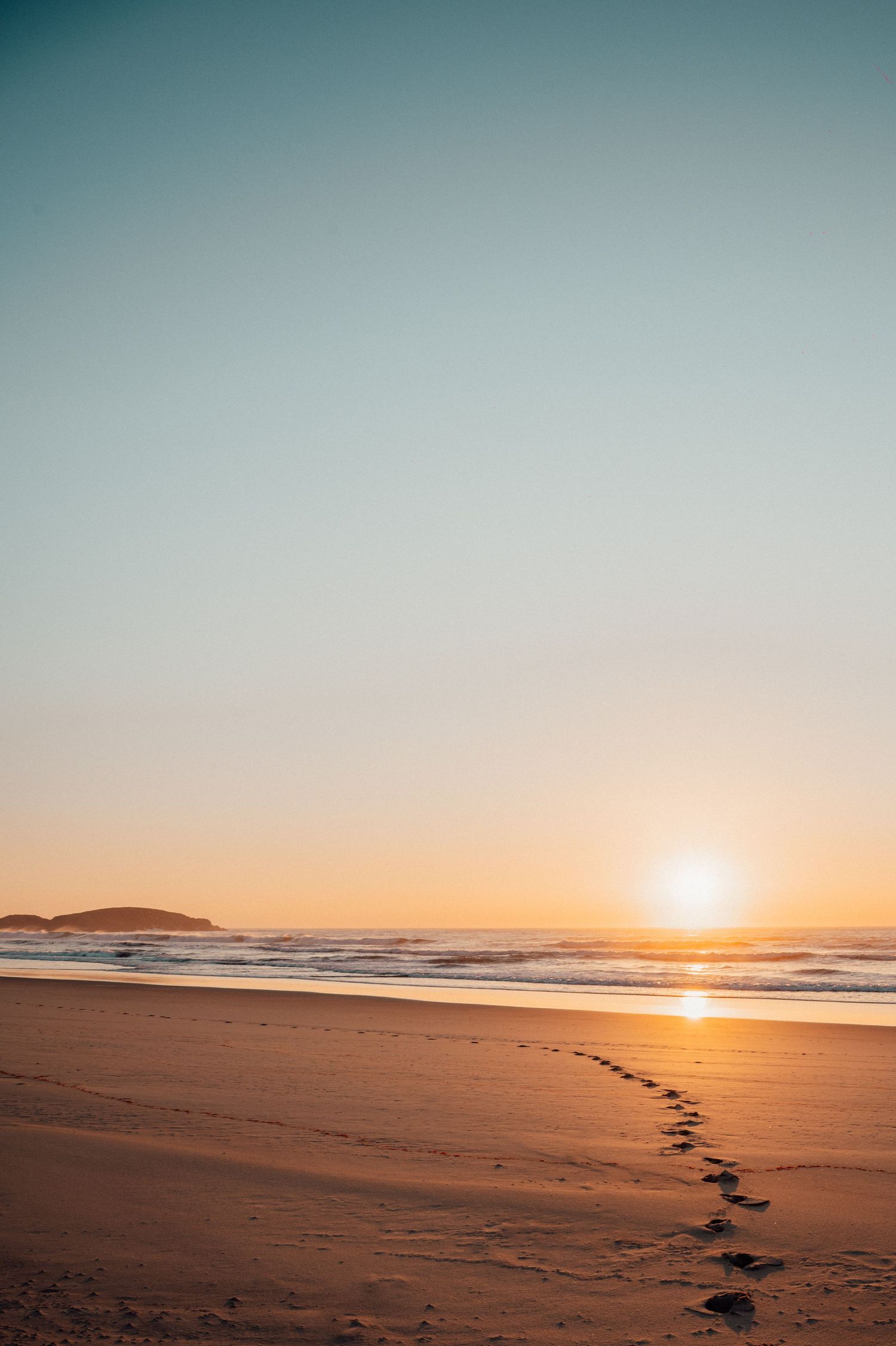 Golden sunset over calm ocean waves gently washing onto a sandy beach with a silhouetted headland on the horizon.