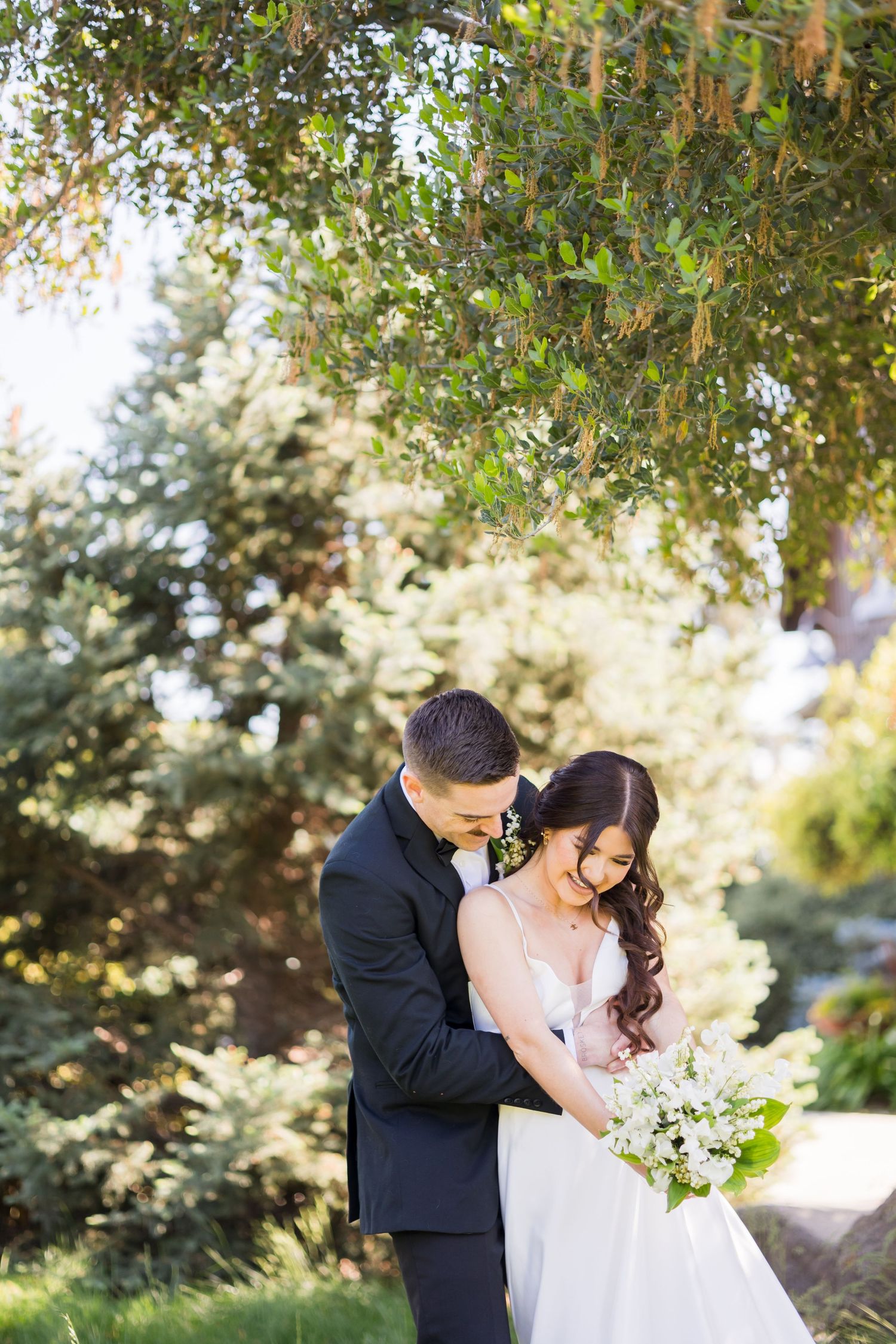 A bride and groom share a tender embrace outdoors, surrounded by lush green trees on their wedding day.