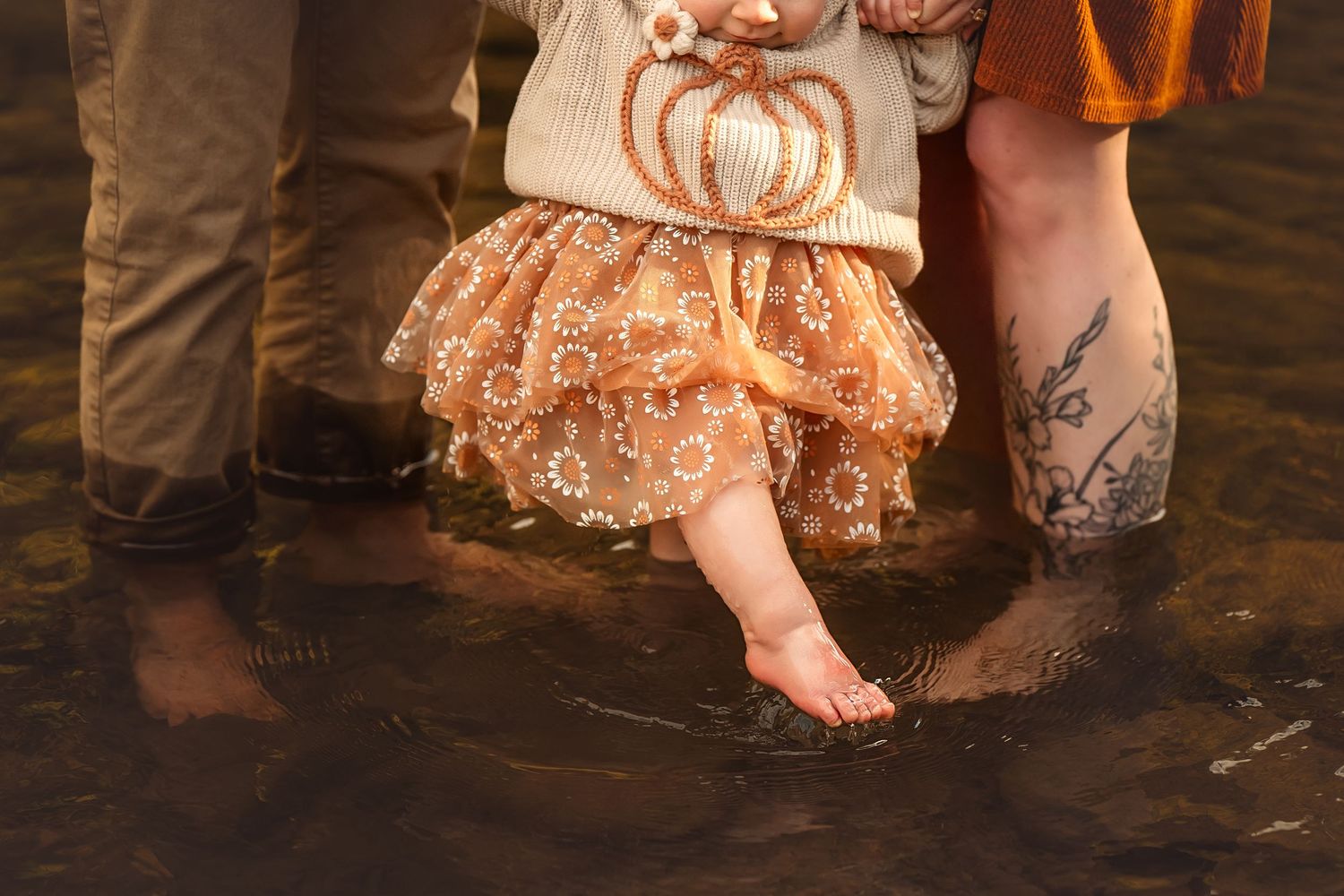 Young girl in sparkly tutu and pumpkin sweater wading in shallow water with adults, fall family photo.
