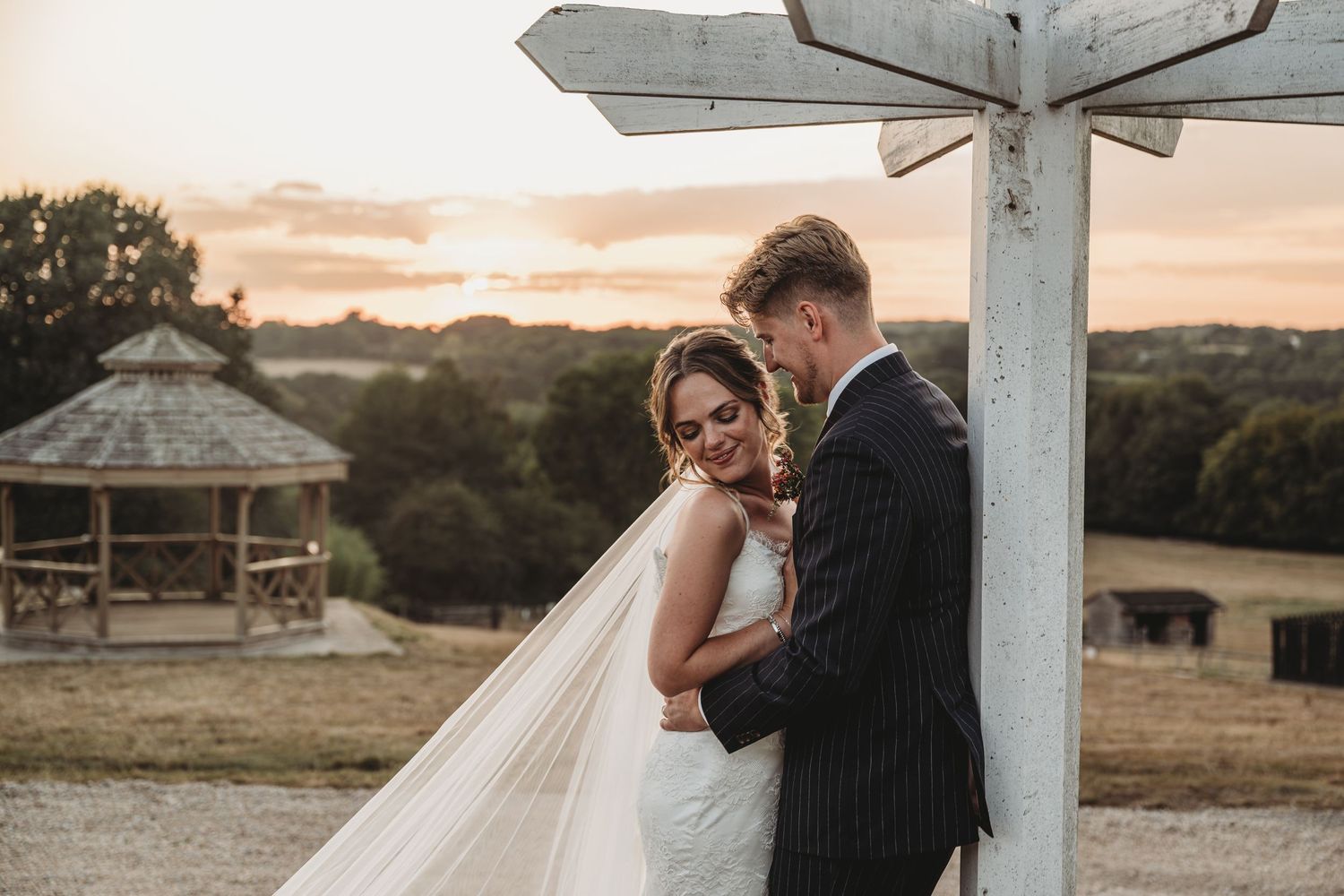 Bride and groom embrace at sunset near a rustic wooden signpost at an outdoor countryside wedding venue.