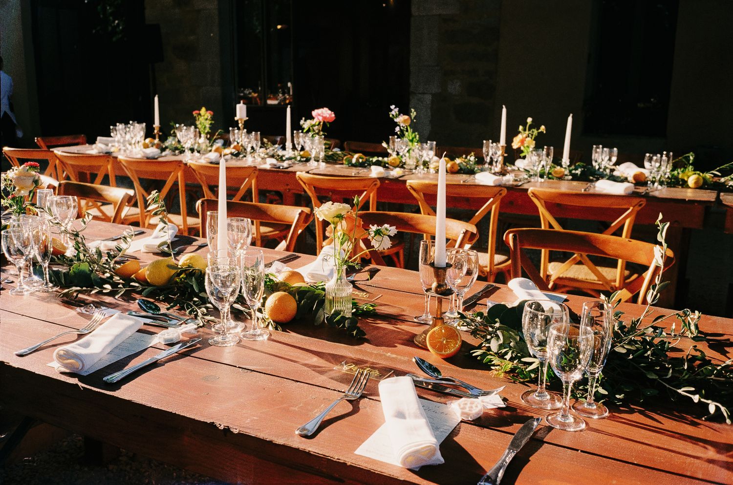 Elegant rustic wedding reception table with greenery, candles, citrus centerpieces, and crystal glassware on a wooden farm table.