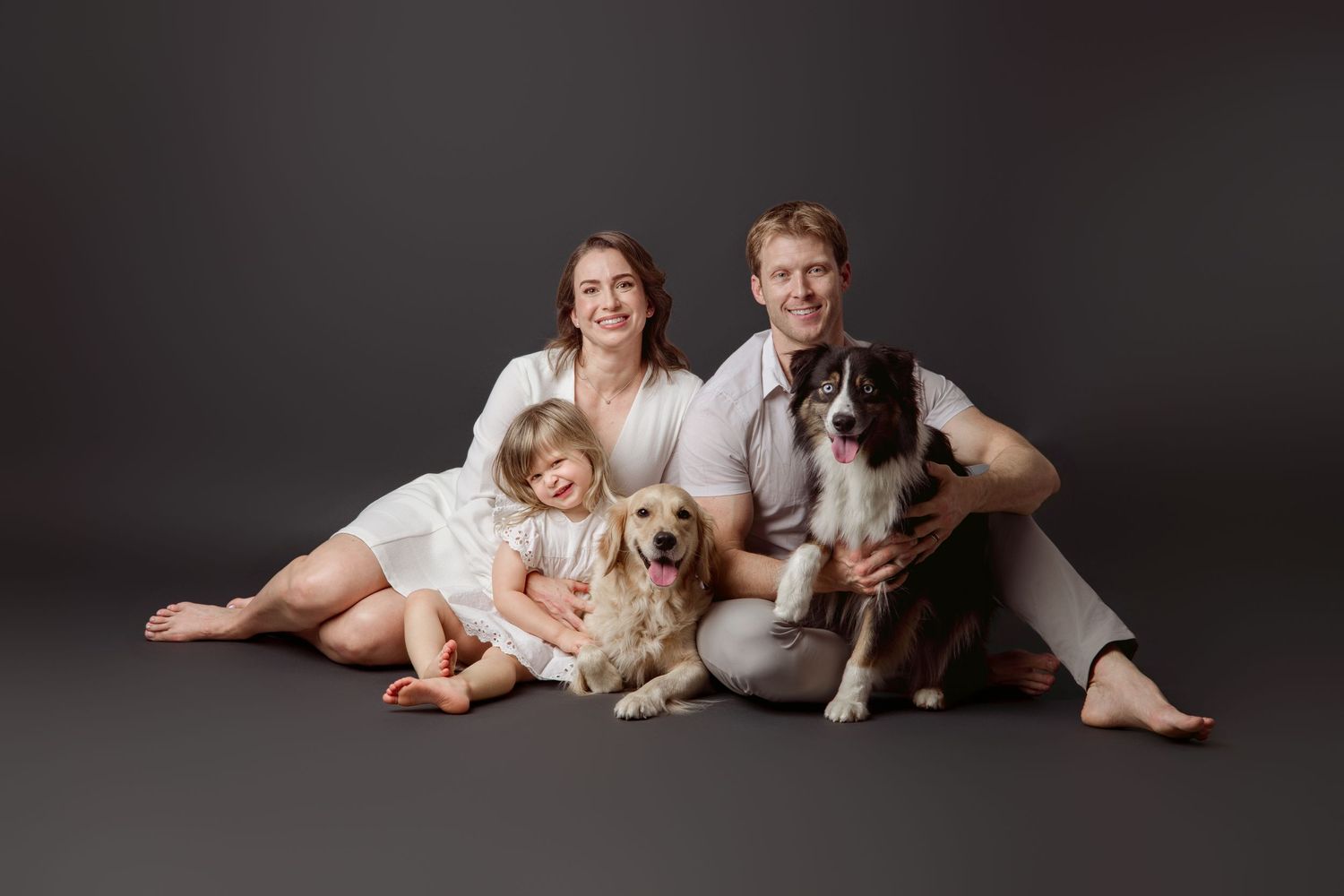 Parents, young daughter, and two dogs seated together in a dark studio