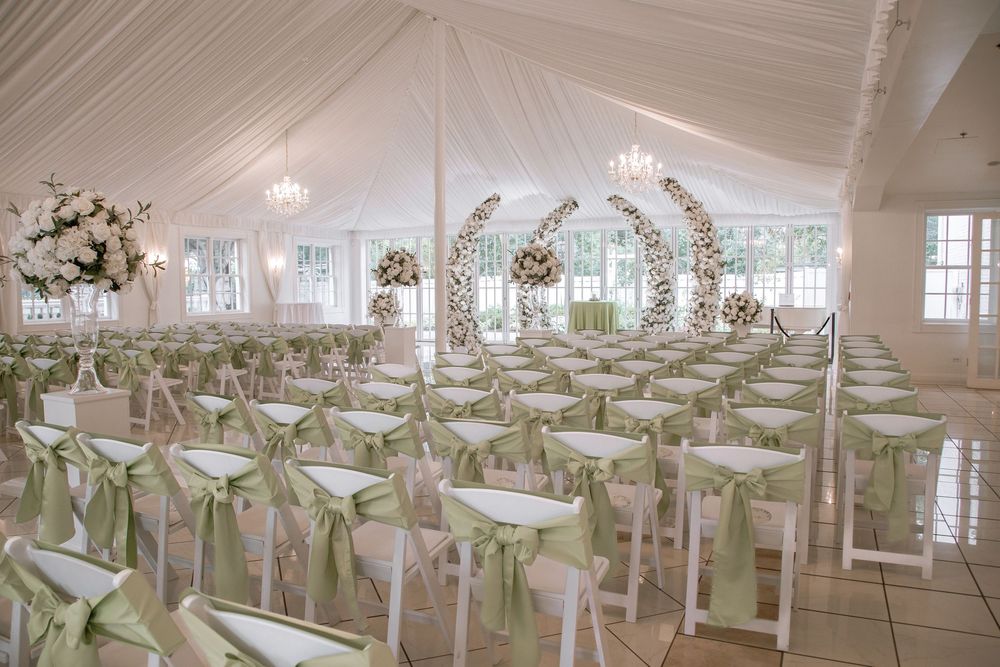 Elegant wedding ceremony tent with white chairs adorned with sage green sashes and floral arch backdrop.