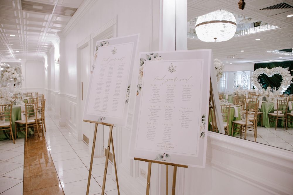 Elegant wedding reception seating chart displays on gold easels in a white ballroom with green chair sashes.