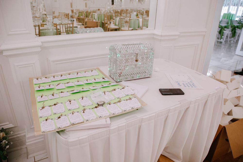 Elegant wedding reception table with white linen, green-accented escort cards, wicker card box, and phone on display.