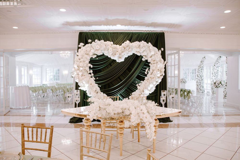 Elegant wedding venue with white floral heart-shaped arch, gold chairs, and dark green backdrop for a romantic ceremony.